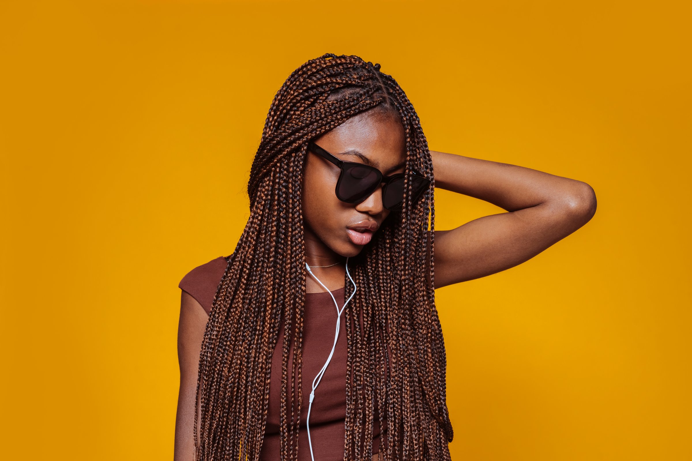 Young african stylish girl with braids and sunglasses, listens music with earphones, dancing in studio isolated over yellow background.