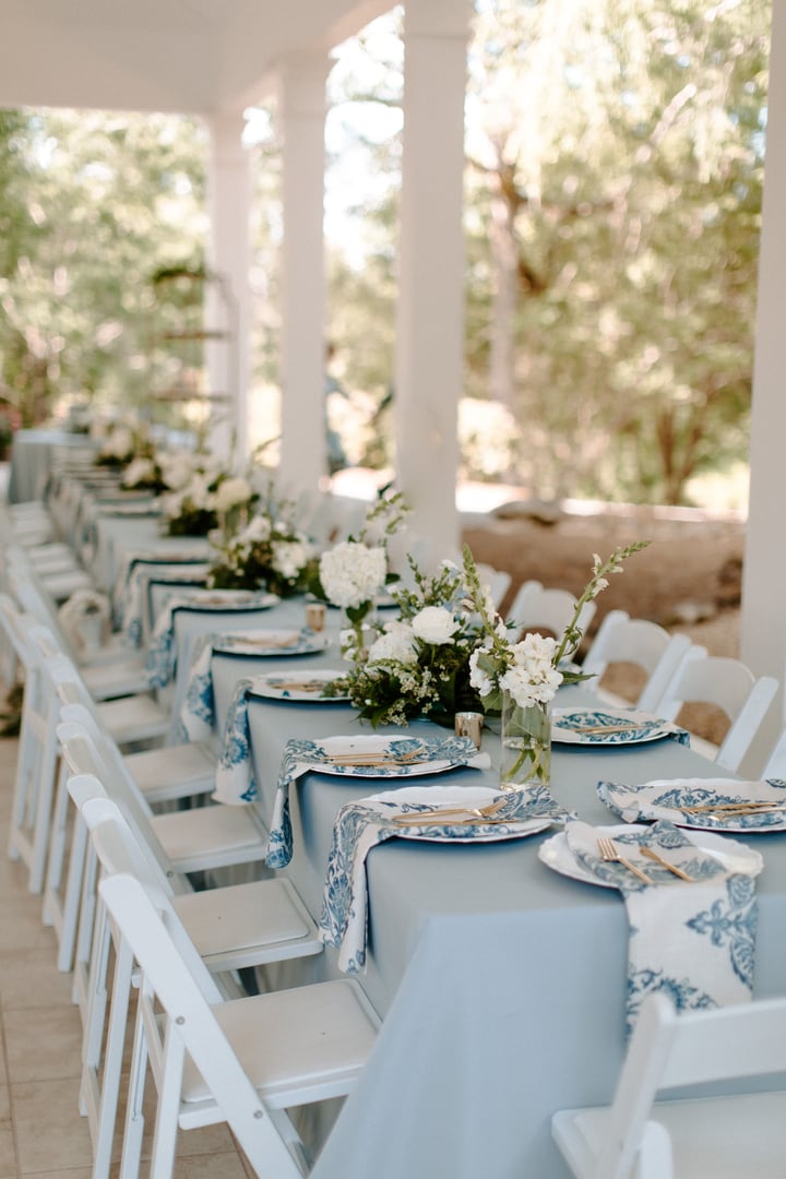 Elegant outdoor wedding reception table setting with floral centerpieces and blue accents under a veranda.