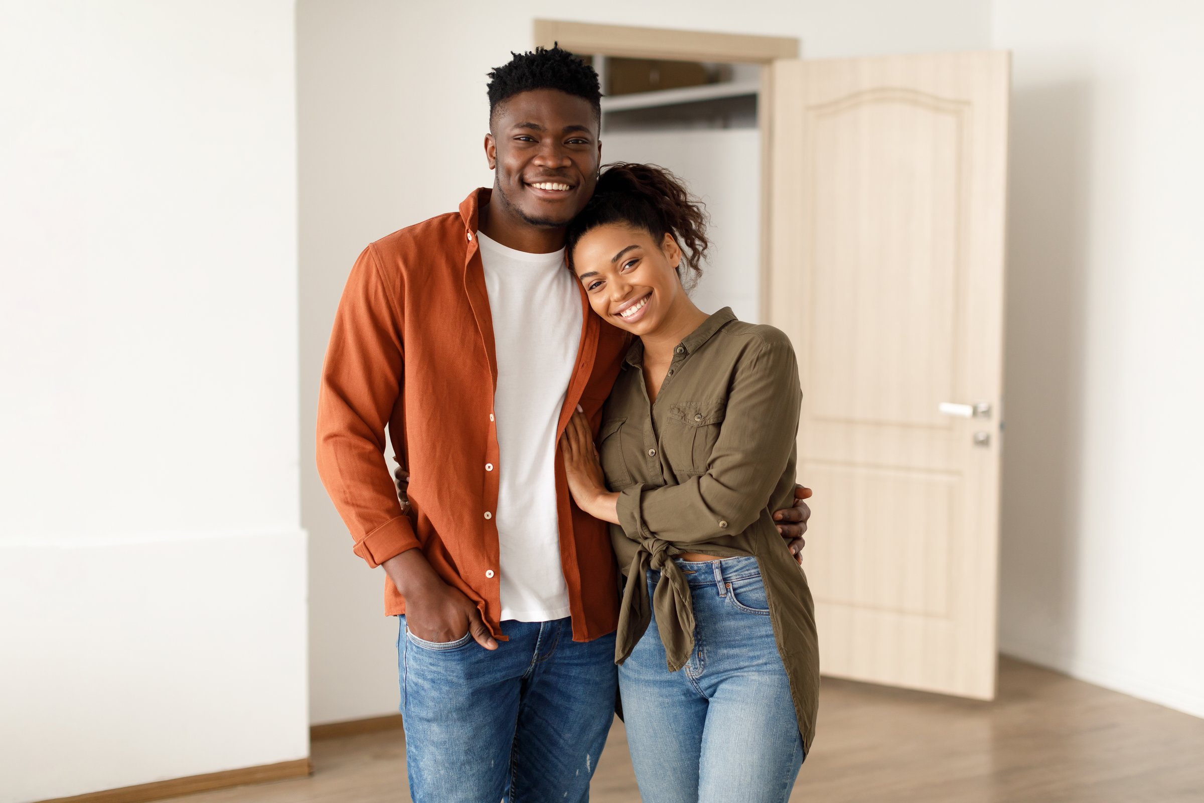 Family Housing. Loving Black Couple Embracing Smiling To Camera Near Opened Door In New Apartment After Relocation. Real Estate Buyers Posing Expressing Happiness And Joy Embracing Inside New House