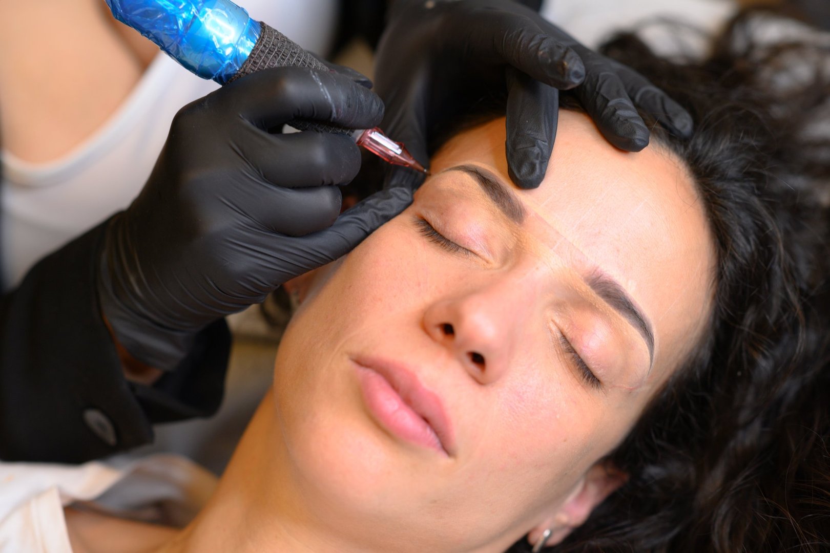 Beautician wearing black gloves applying permanent makeup on eyebrows of a brunette woman lying down with her eyes closed