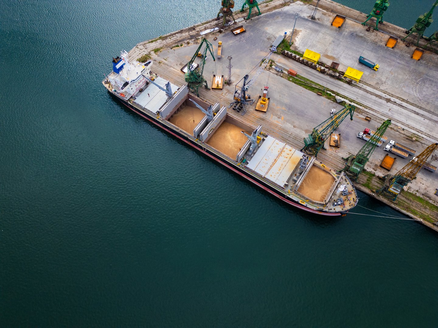 The cargo ship, docked at the harbor, is being uploaded as cranes lift grains from the open holds. Surrounding machinery and equipment are visible.