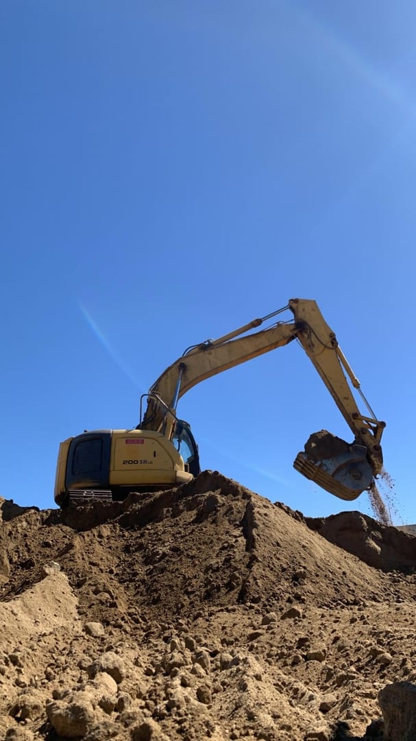 Excavator on a dirt mound under clear blue sky, lifting soil with its bucket, in a construction or mining scene.