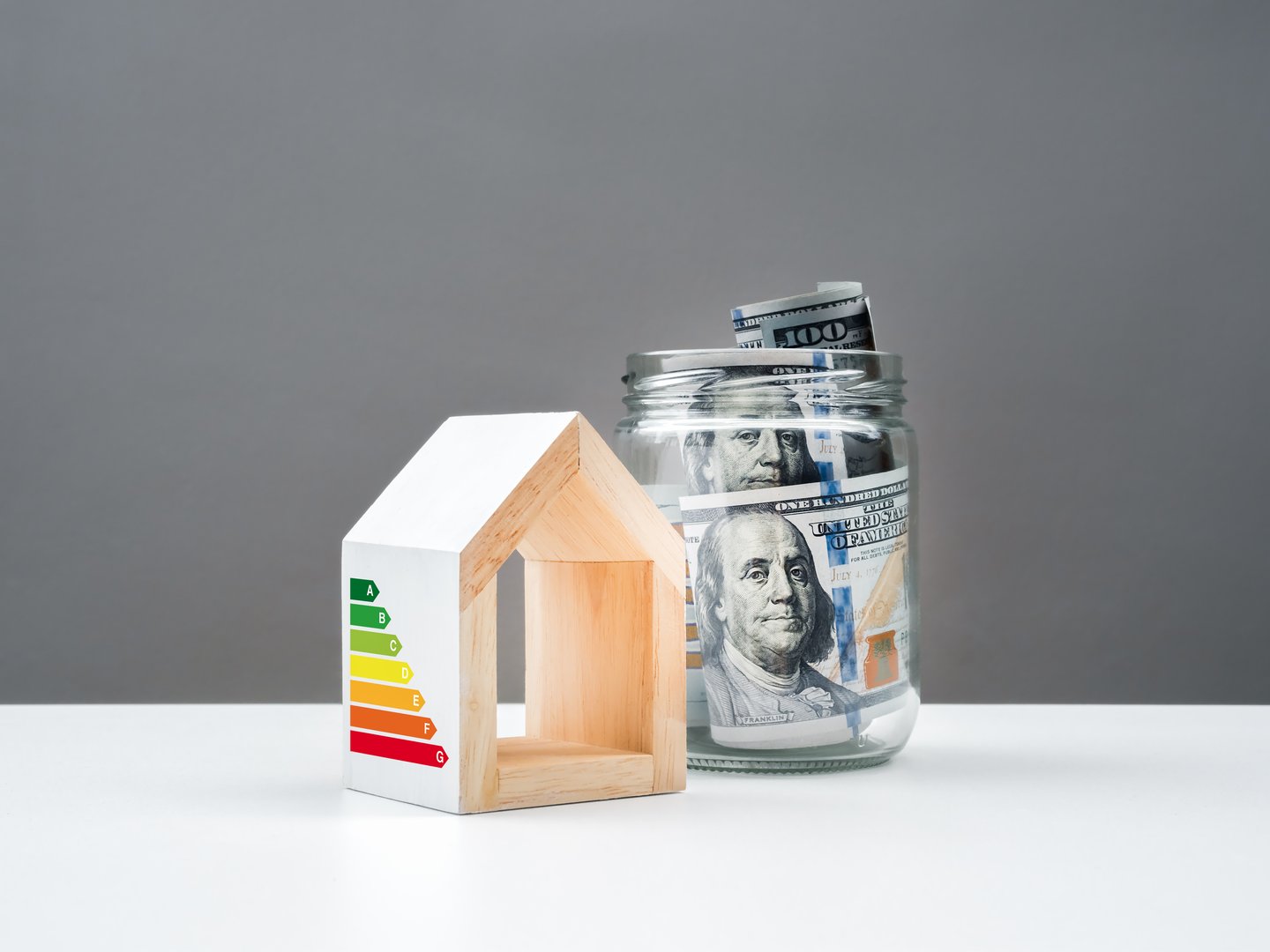 Efficient energy concept, sustainable energy efficiency saving home. One hundred dollars bill banknotes in glass jar near white house model with rate label audit on white table, grey background.