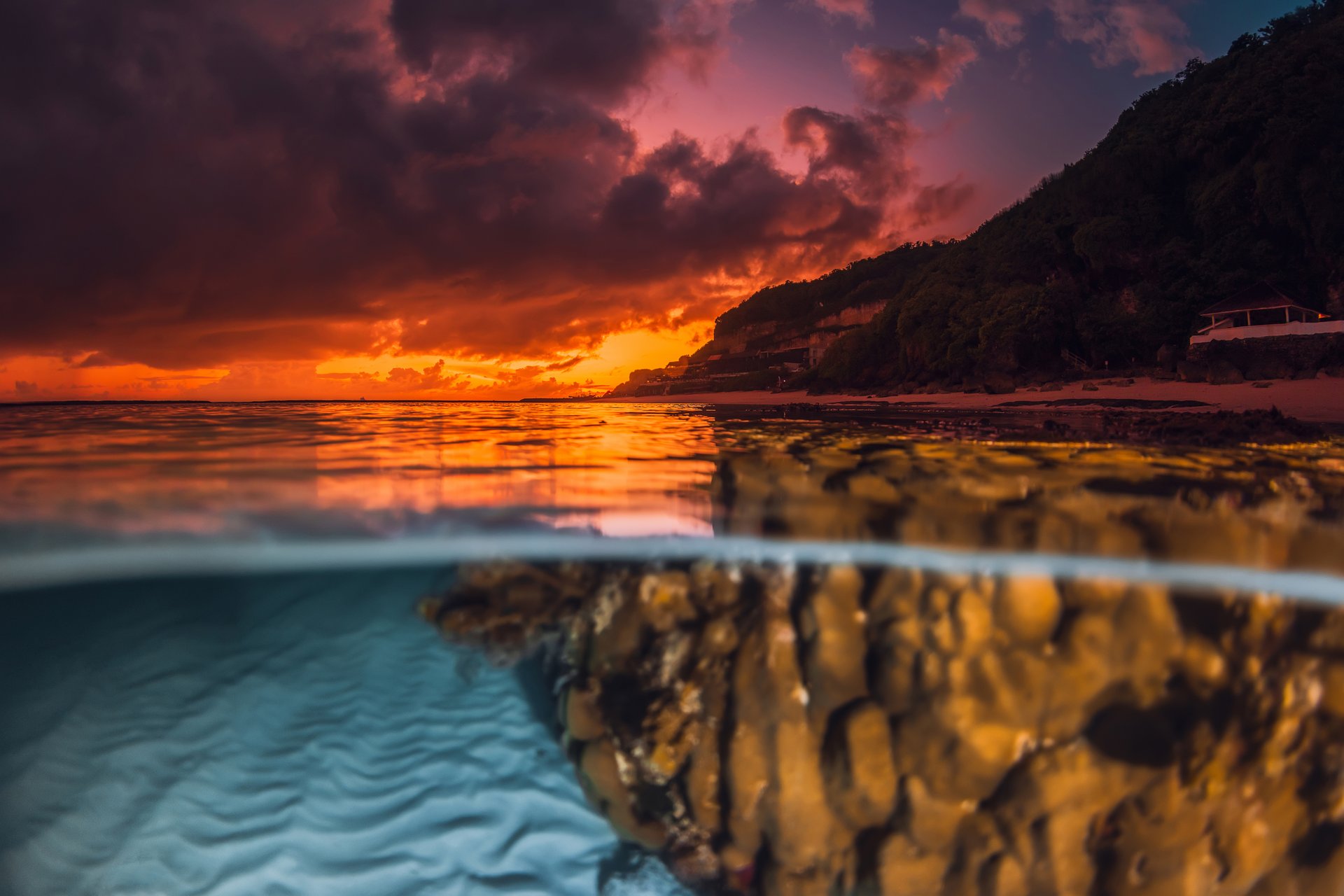 Dramatic sunset or sunrise over calm sea with underwater sand patterns and coral, captured in a split view seascape
