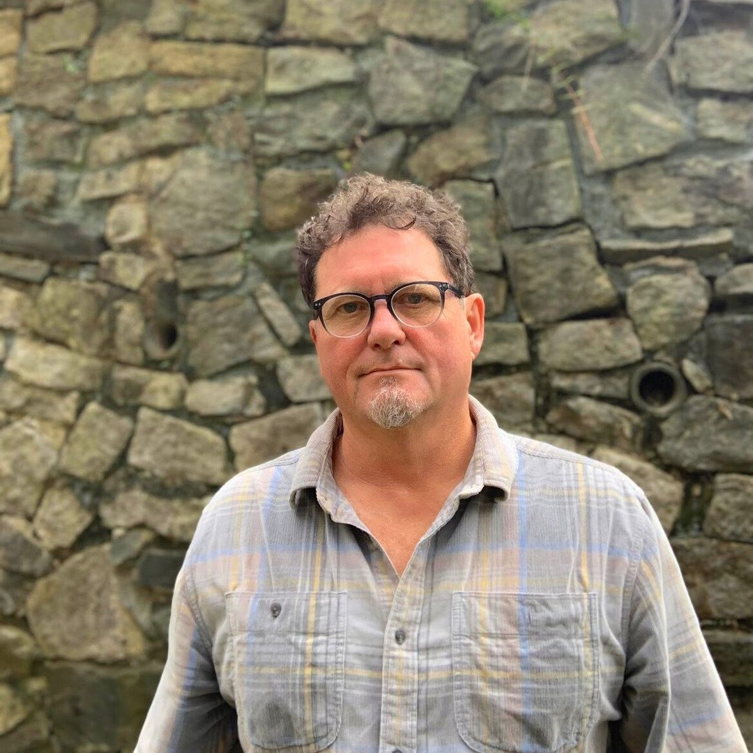 Man with glasses and a plaid shirt standing in front of a stone wall, looking at the camera.