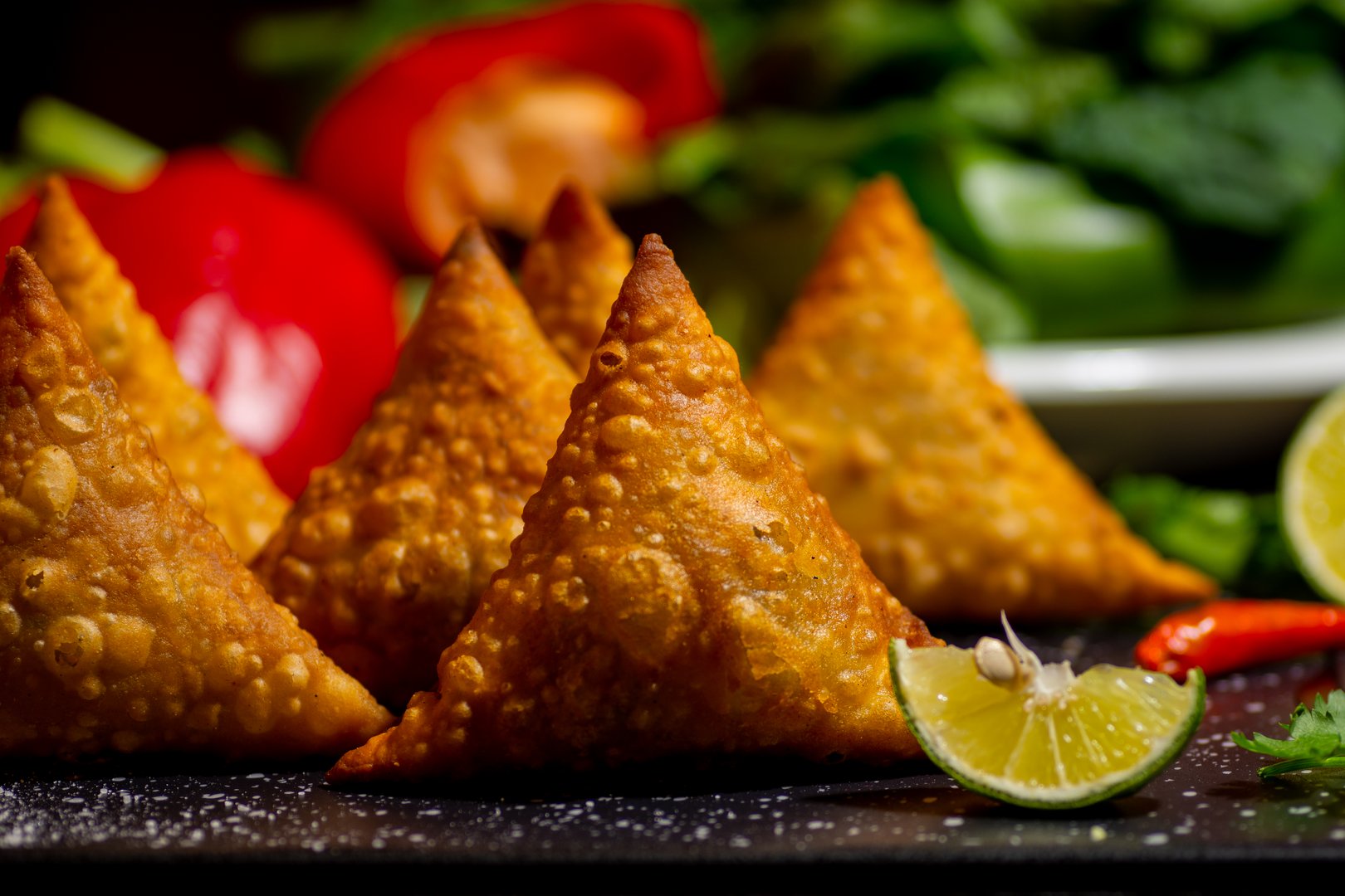 Close-up of crispy samosas with lime wedges, surrounded by fresh vegetables in the background.