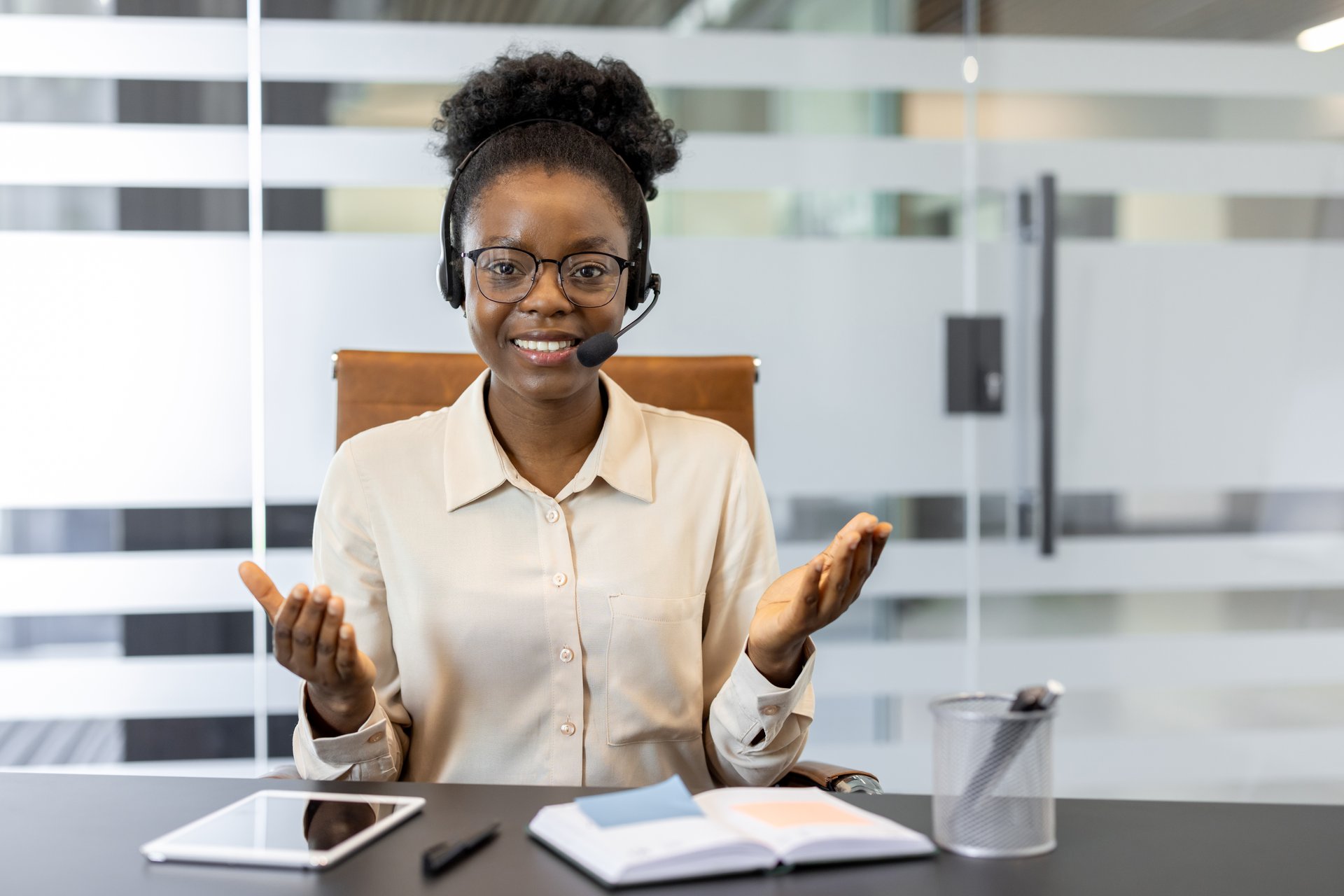 Smiling customer service representative wears headset in modern office. Engages in video call using phone, showing friendly approach. Display of open notebook and digital tablet