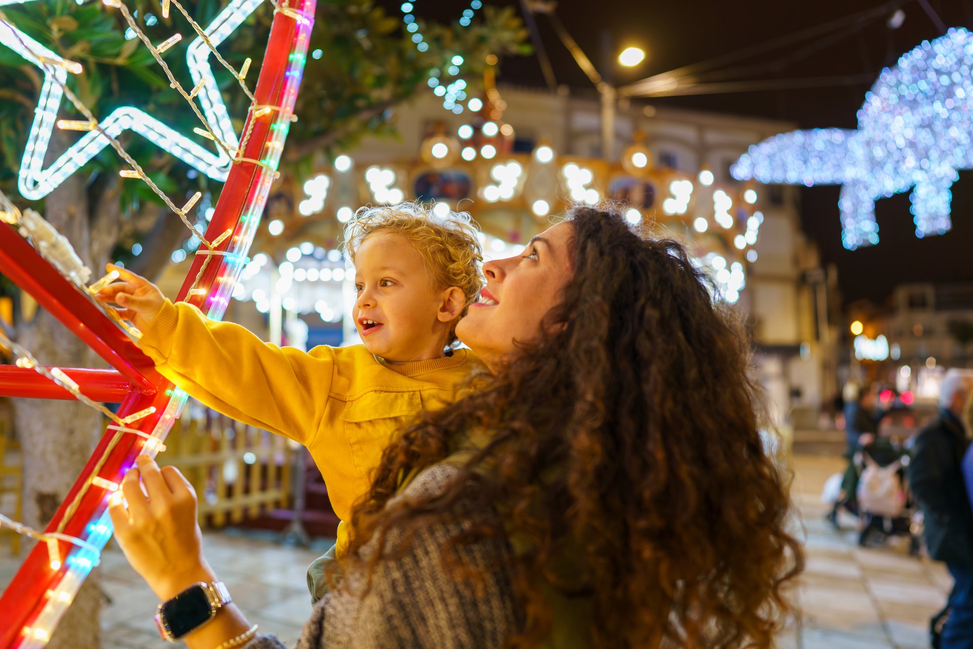 Mother carrying her son, enjoying christmas lights at a festive night market, filled with warmth and joy