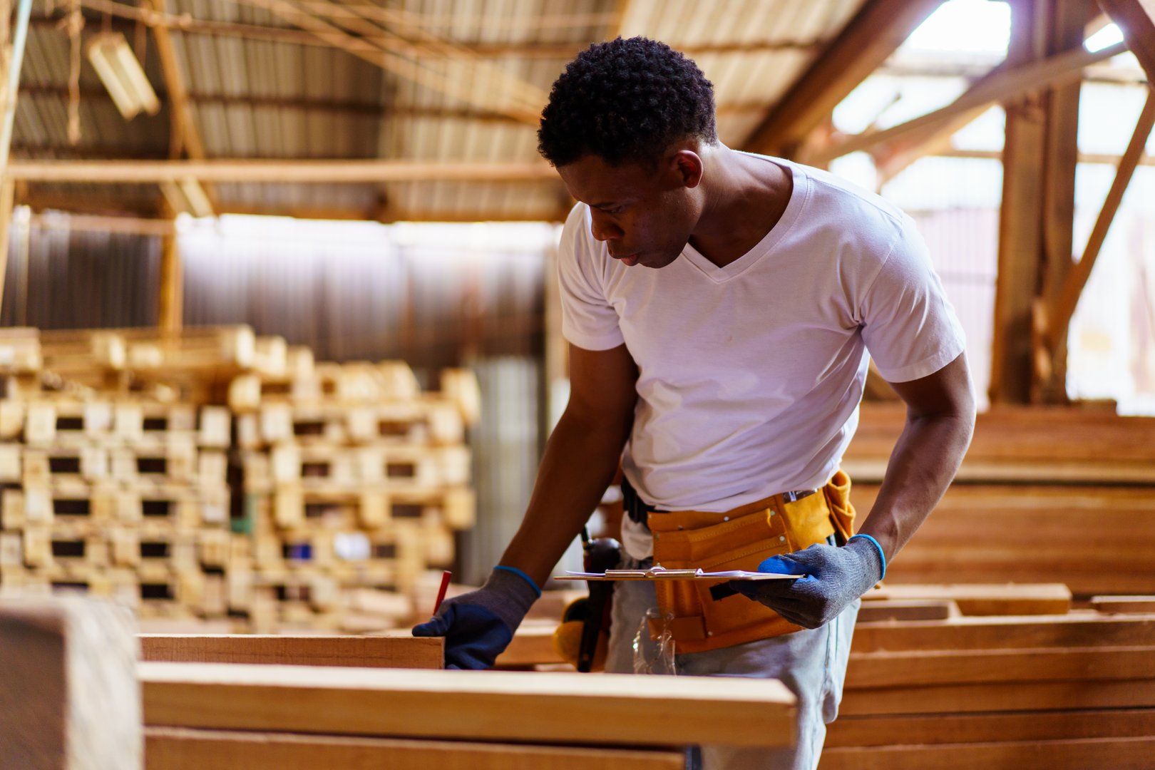 African American carpenter inspecting wood and timber in a sawmill, holding documents, checking quality of lumber production, working with professional skills in woodworking and carpentry industry.