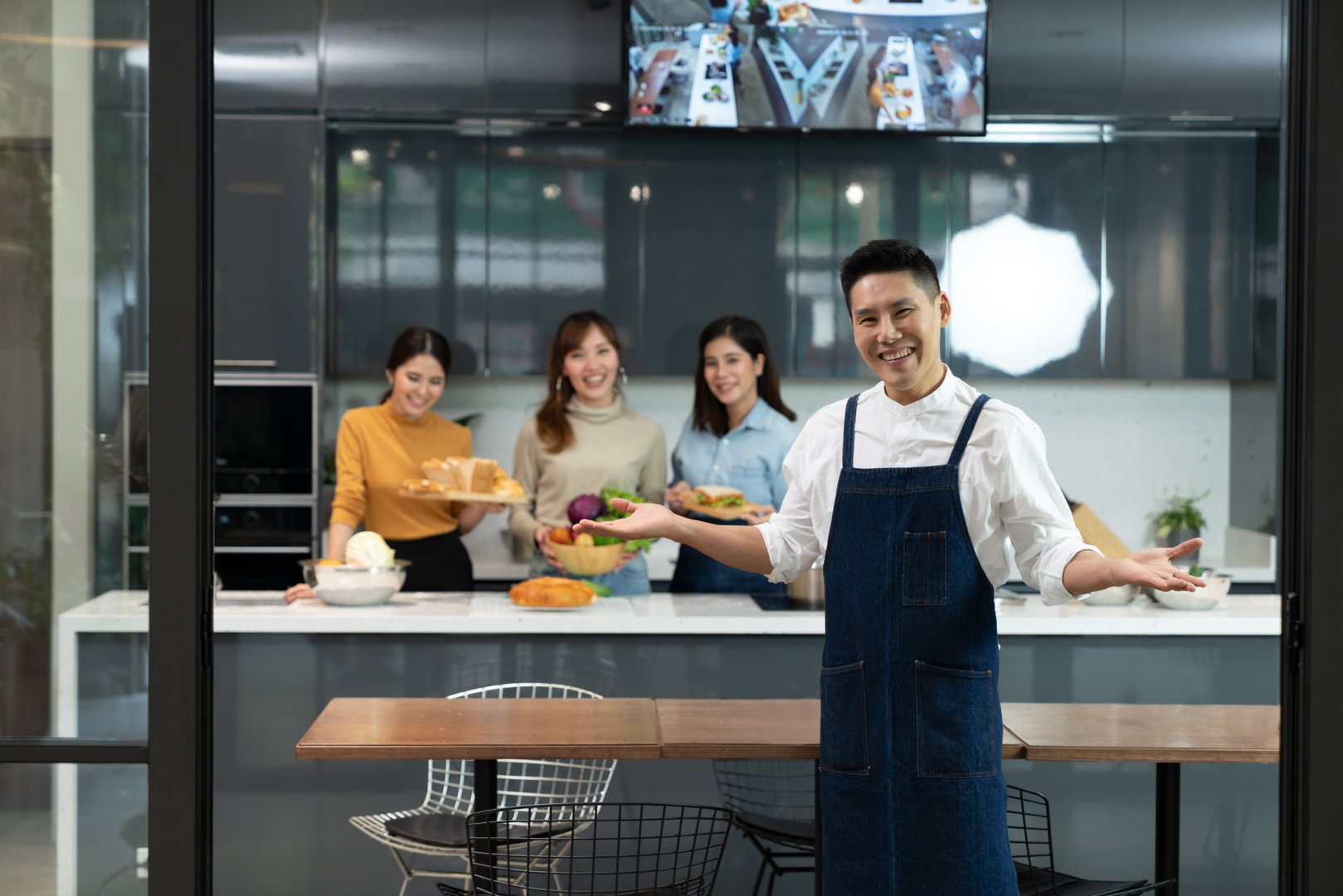 Asian executive master chef in blue apron posing invite to enjoy a private group cooking class by standing in front of trainee student chef background. Concept of culinary course and Kitchen school.
