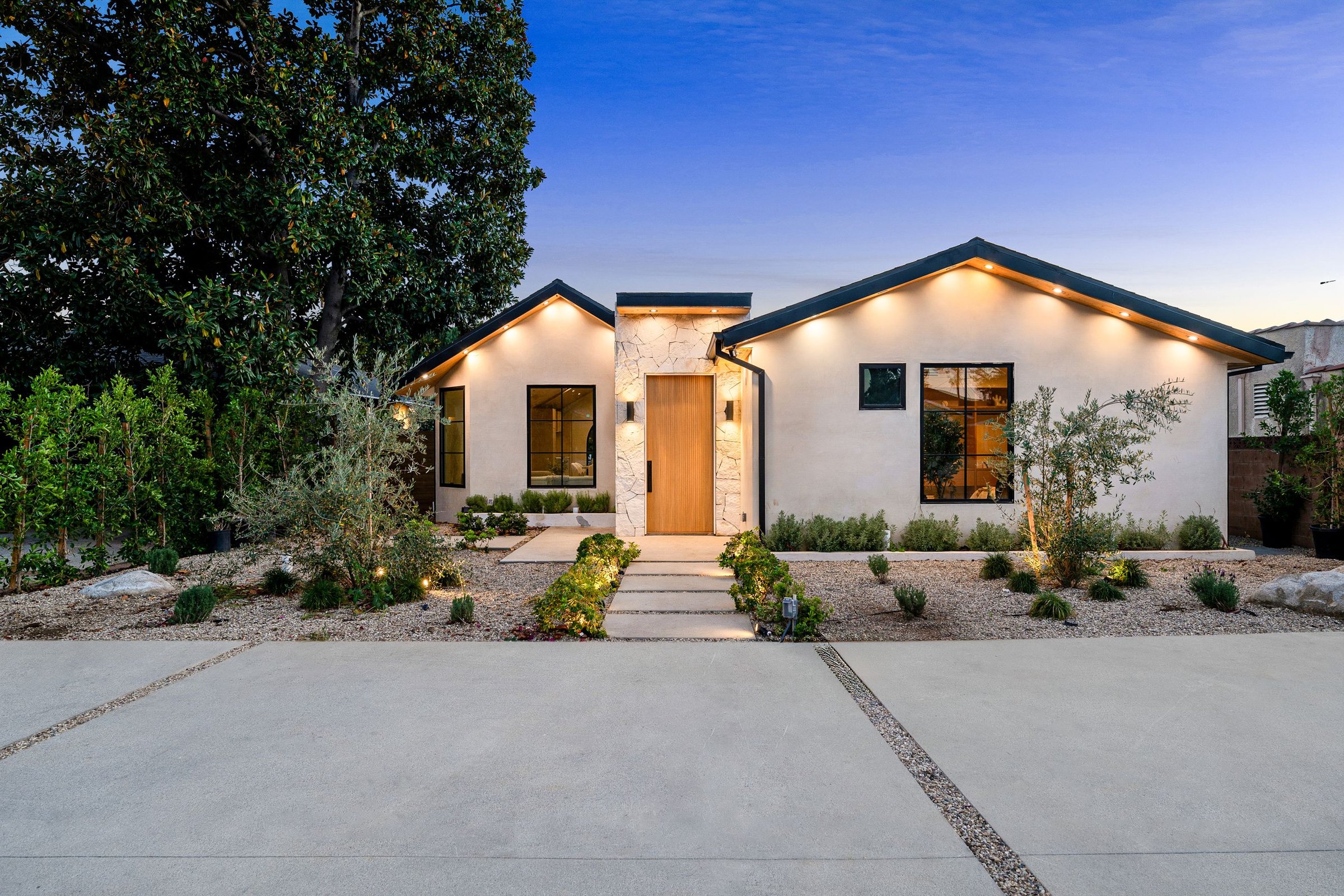 Modern single-story house with landscaped front yard, illuminated at dusk, featuring a minimalist design and natural materials.