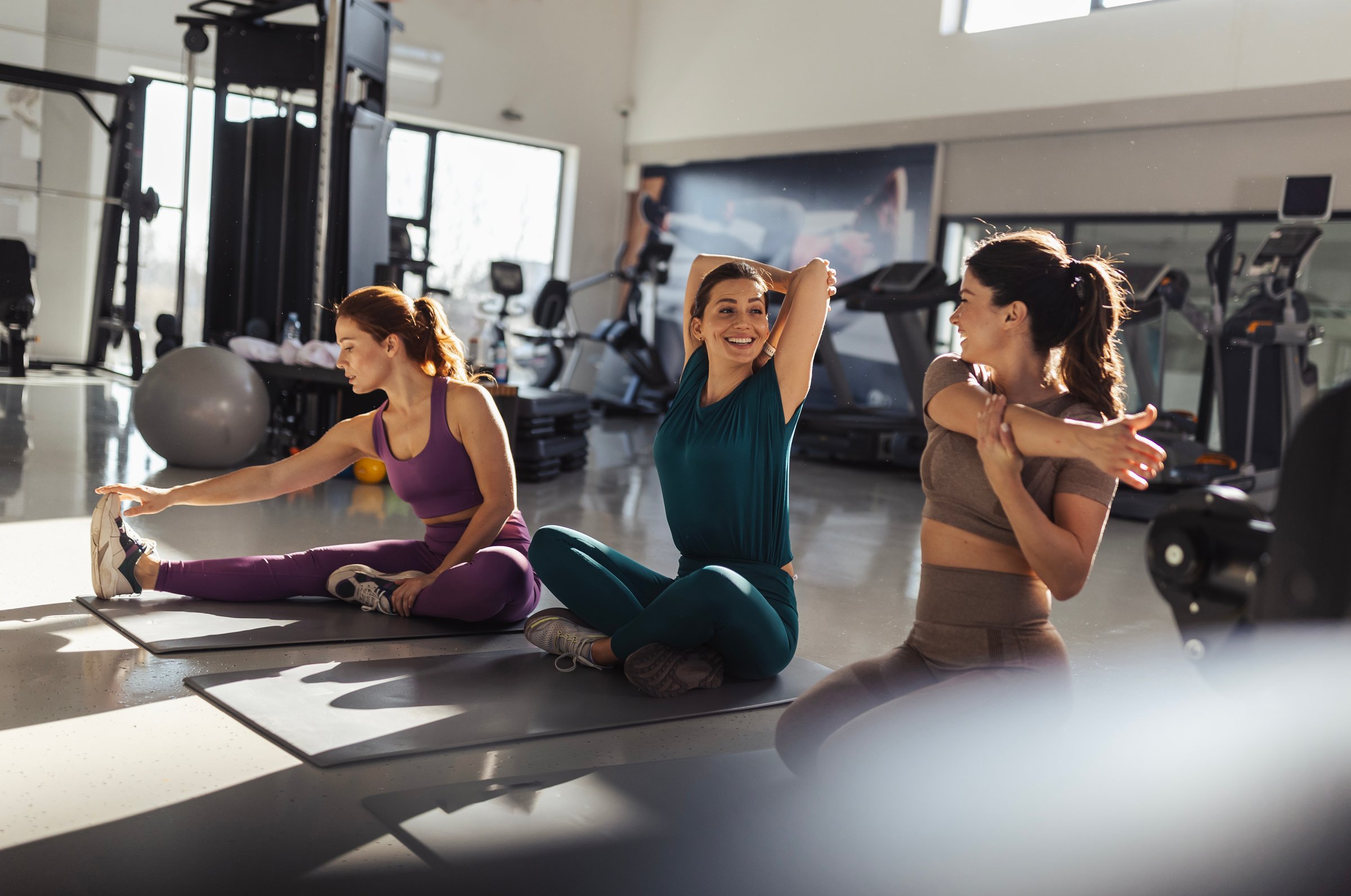 Three young women are stretching on the gym floor, engaging in warm-up exercises. They are smiling and enjoying their time together in a fitness environment
