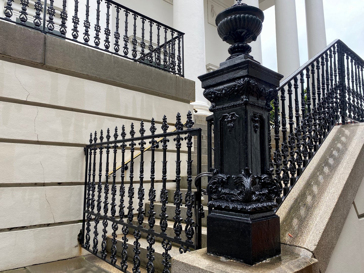 An elegant cast-iron stair railing and column post on a historic building in Savannah, Georgia. The black wrought iron contrasts with the pale stone exterior, highlighting the city's architectural craftsmanship and Southern sophistication.