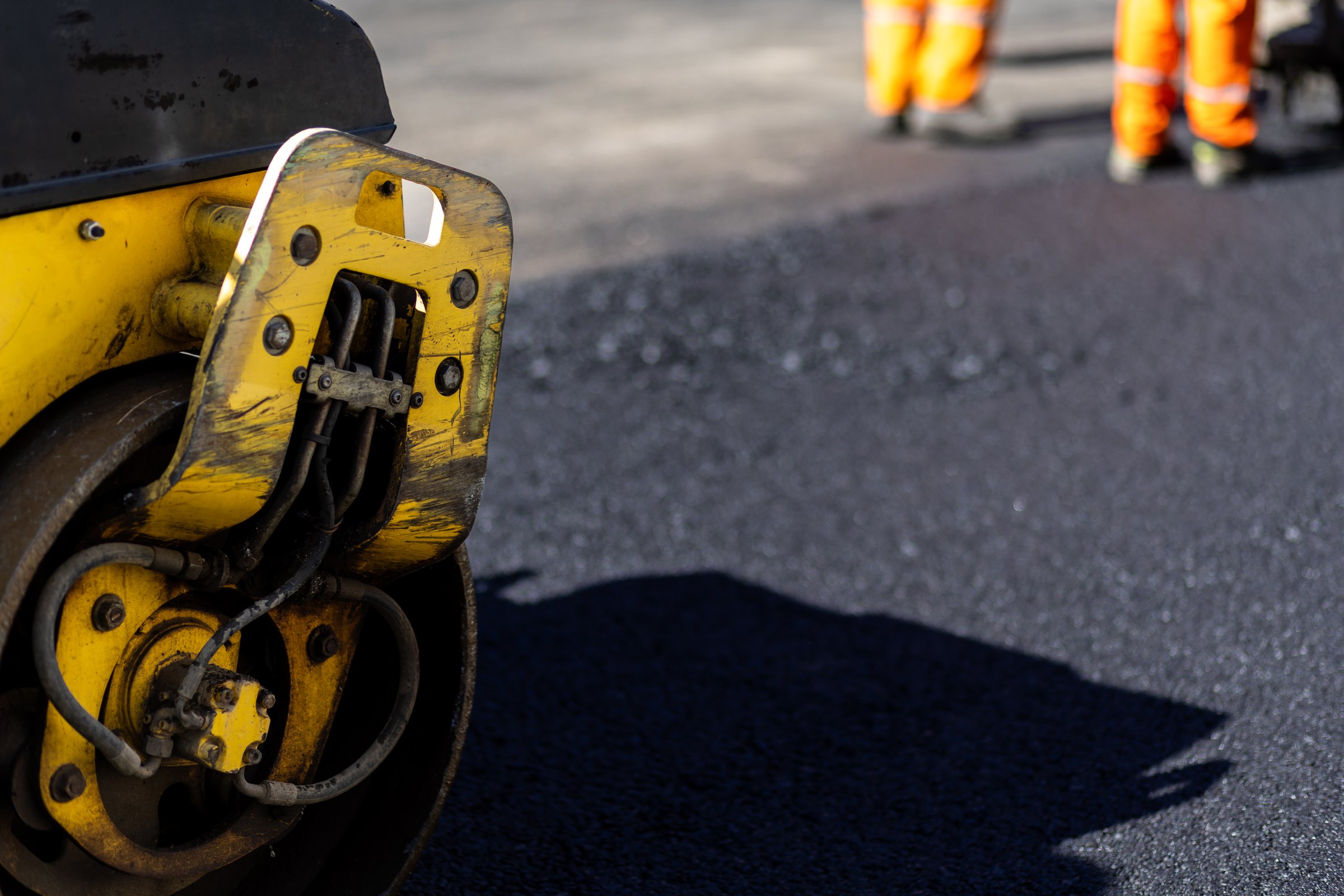 A road roller is being used to smooth freshly laid asphalt, while construction workers in high-visibility gear are involved in the resurfacing project.