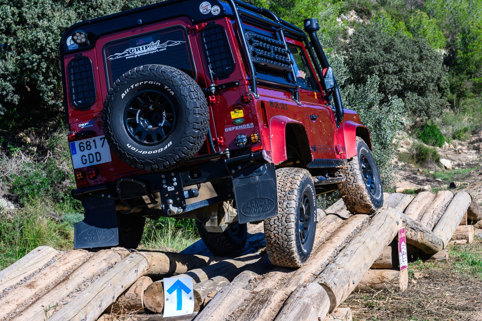 Land Party 2024, Les Comes, Suria, Barcelona, Spain Dark shiny red Land Rover Defender on a wooden bridge-crossing obstacle at an exhibition, with the car tilted and a wheel hanging down. It has a spare wheel mounted on the rear door.