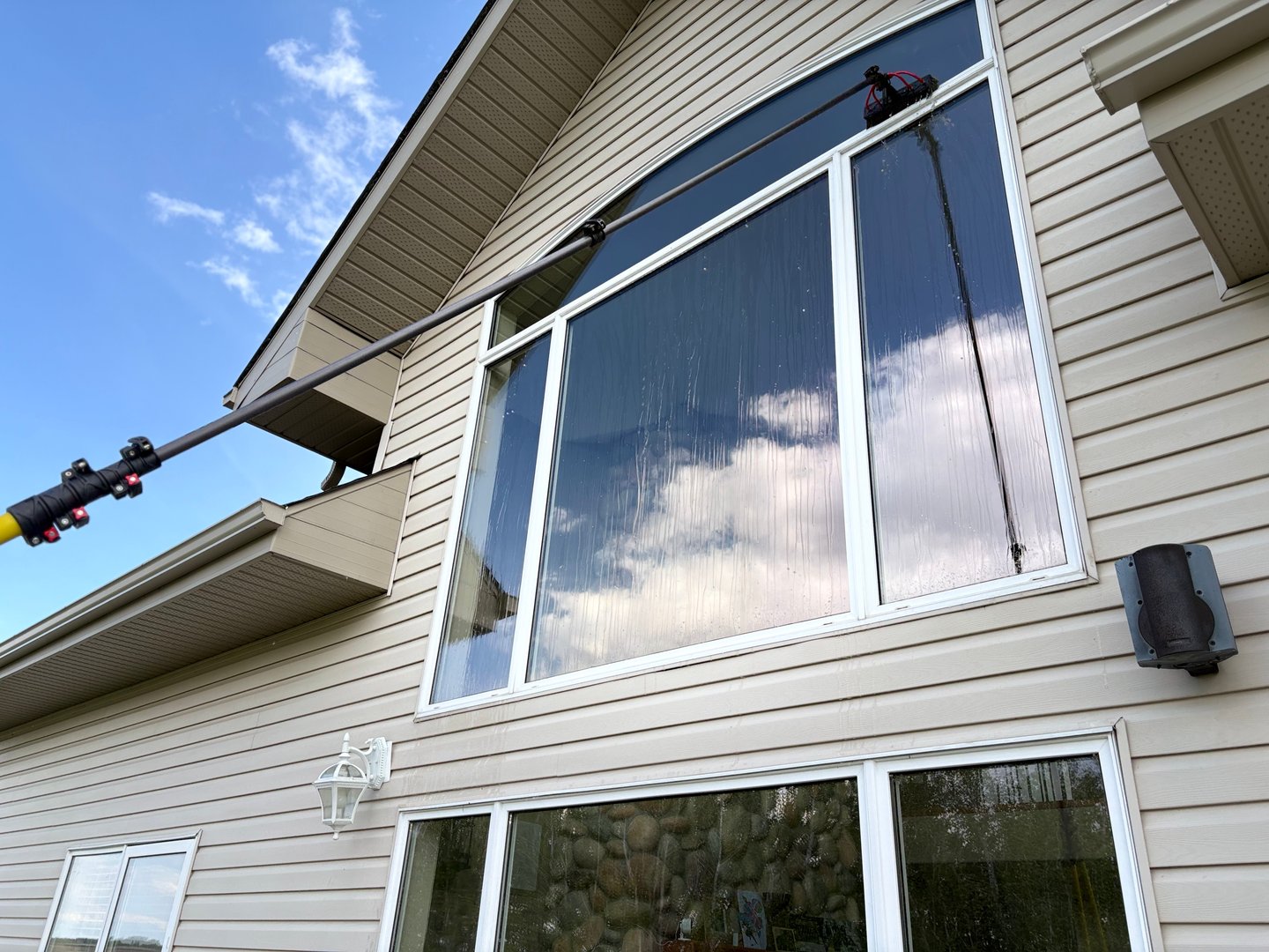 Person cleaning large house windows using an extendable pole on a sunny day with a visible reflection in the glass.