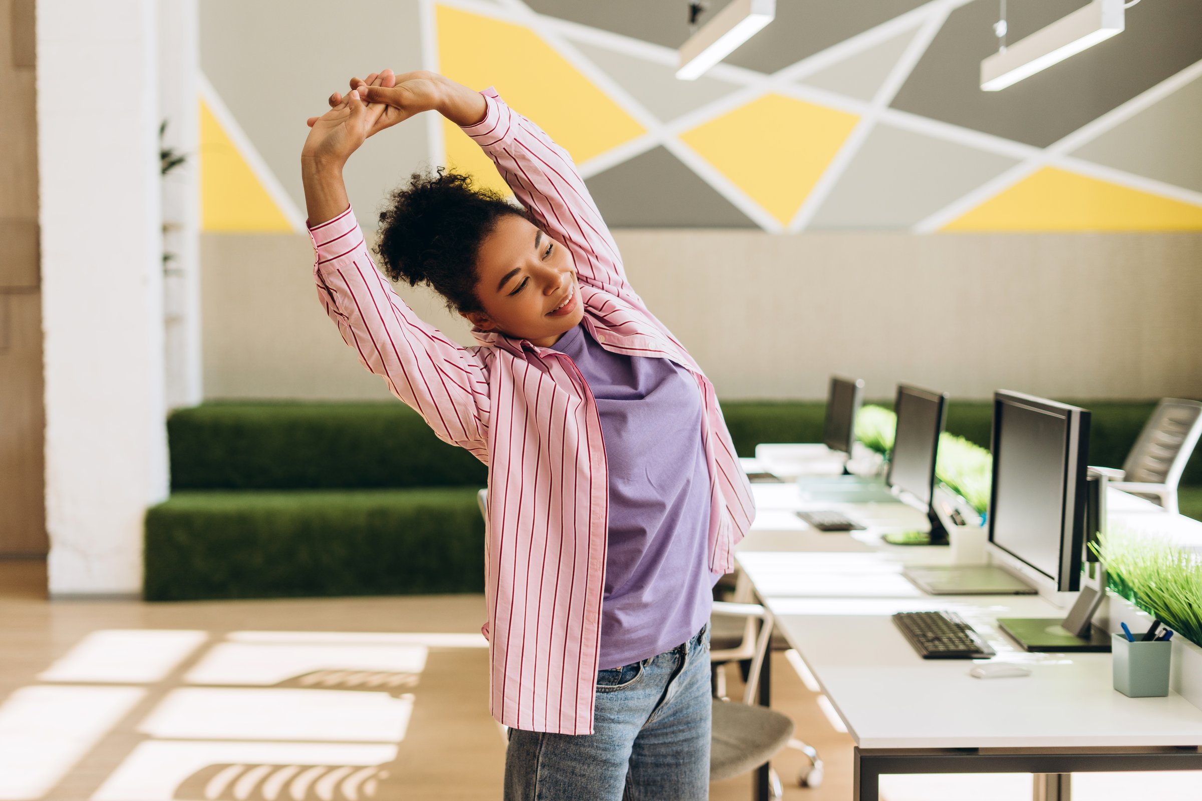 Happy young african american manager woman is stretching her arms while standing near her desk in modern office, enjoying break time during workday