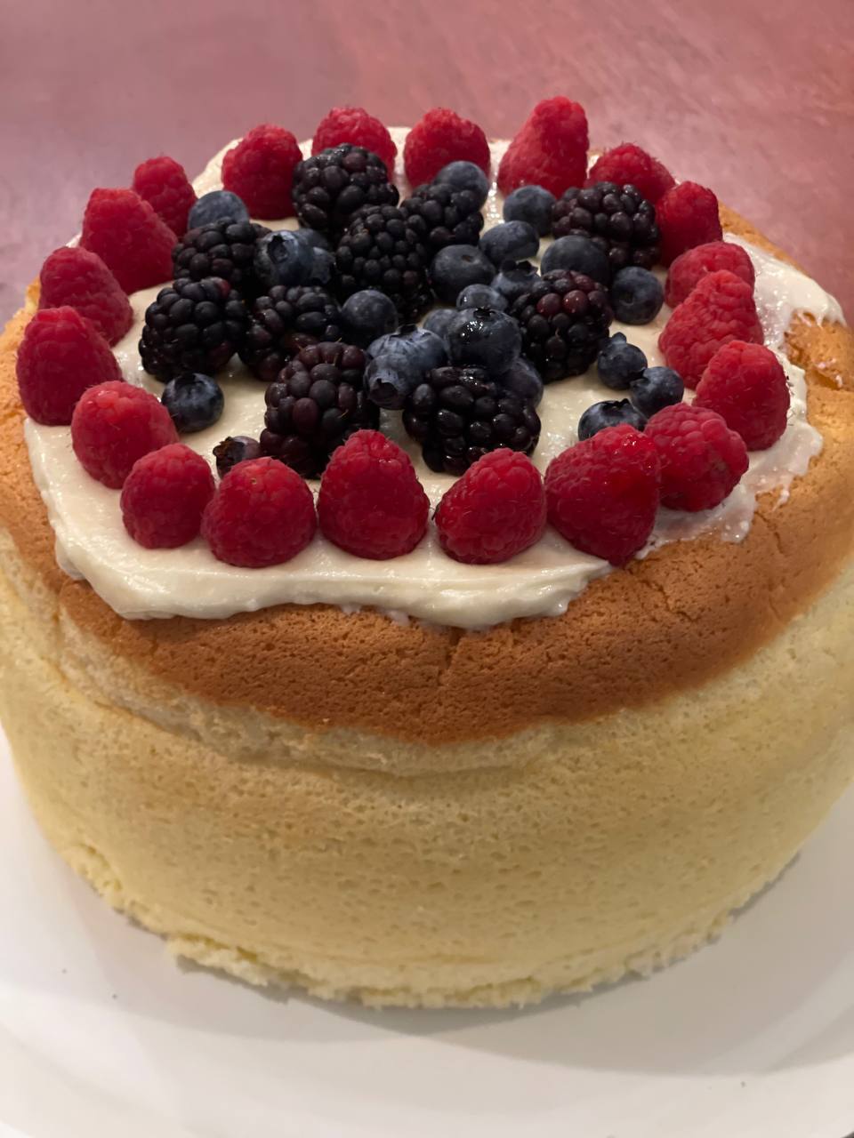 A sponge cake topped with cream, raspberries, blackberries, and blueberries, arranged in a circular pattern.