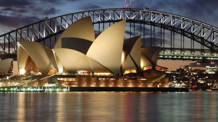 Sydney Opera House at night, illuminated with lights, with Sydney Harbour Bridge in the background.