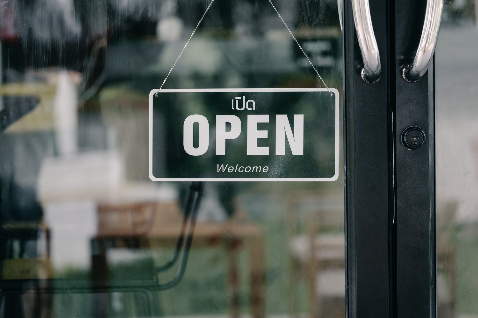 Black open sign with welcome text hanging on glass door of modern cafe, showing business opening hours and customer invitation.