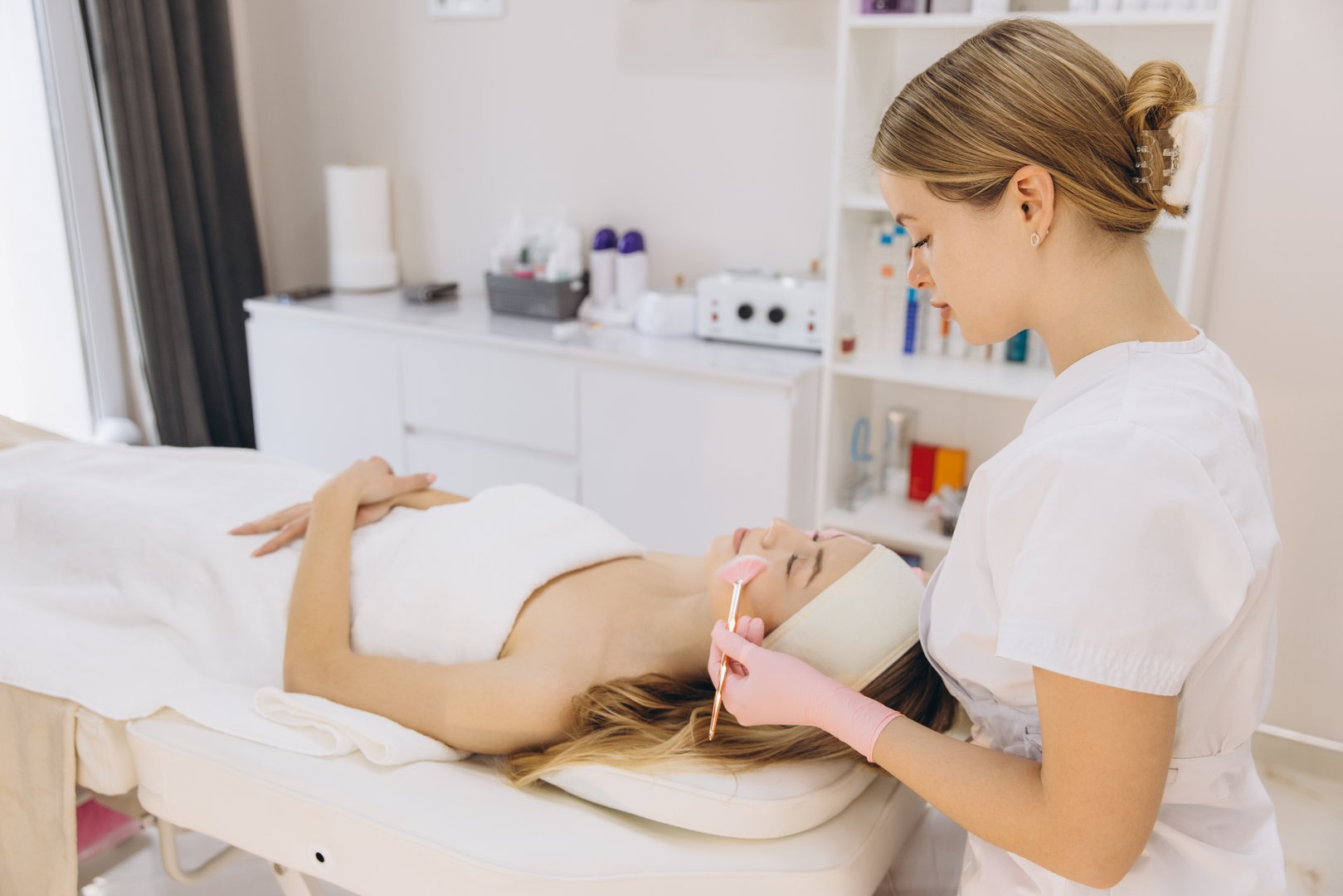 Professional beautician applying a facial treatment to a young woman lying on a bed in a beauty center, skincare and cosmetology concept
