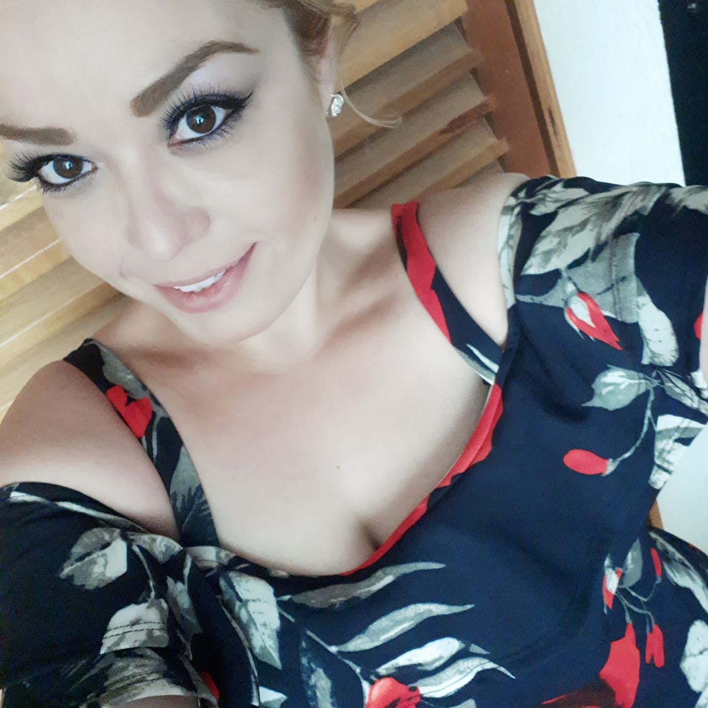 Woman with makeup and floral off-shoulder top smiling and looking at the camera, against a wooden backdrop.