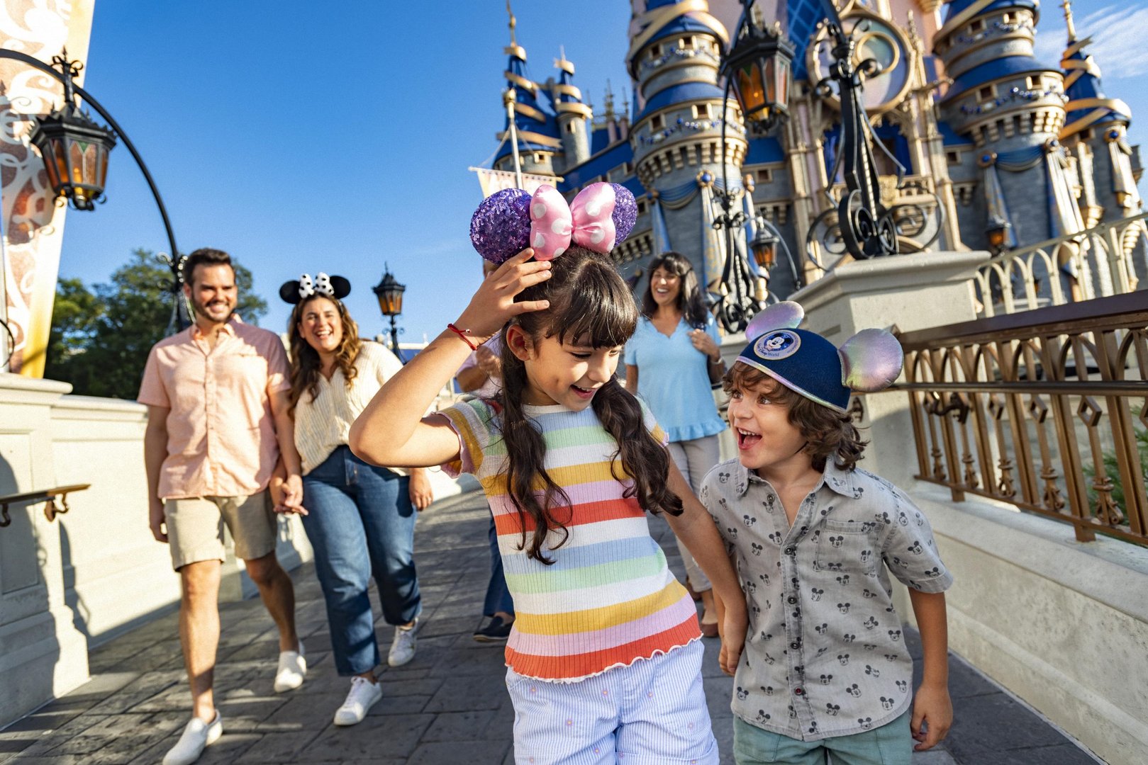 Children wearing Mickey ears laughing in front of a castle, with adults walking behind them in a theme park.