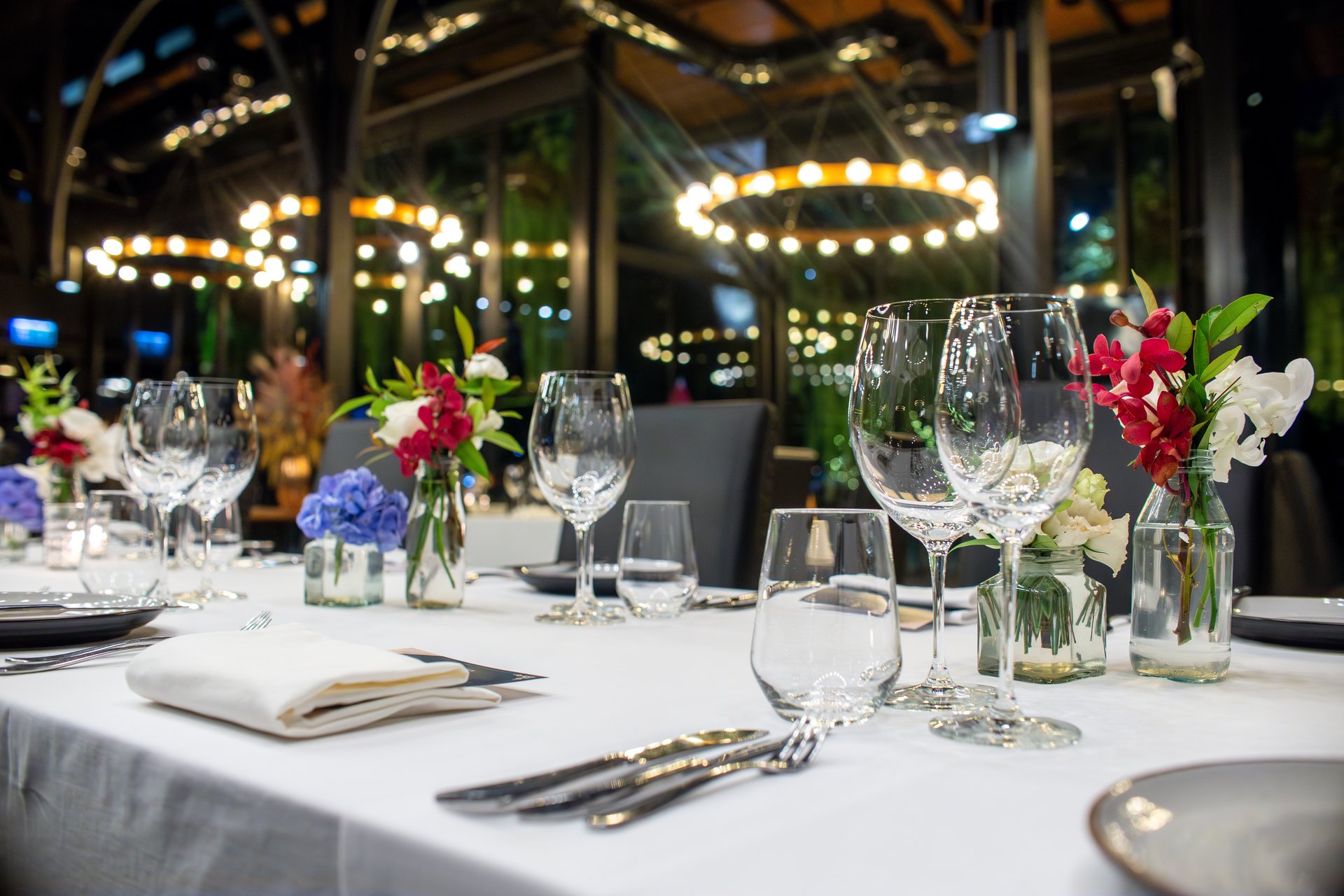 A luxurious dinner table setup in a modern venue with grand lighting and floor-to-ceiling windows. Floral centerpieces and polished glassware enhance the upscale, sophisticated atmosphere.