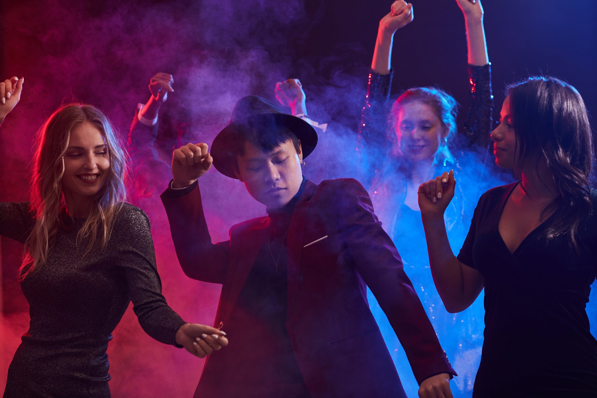 People dancing energetically in a club with colorful lights and smoke effects, wearing party attire.