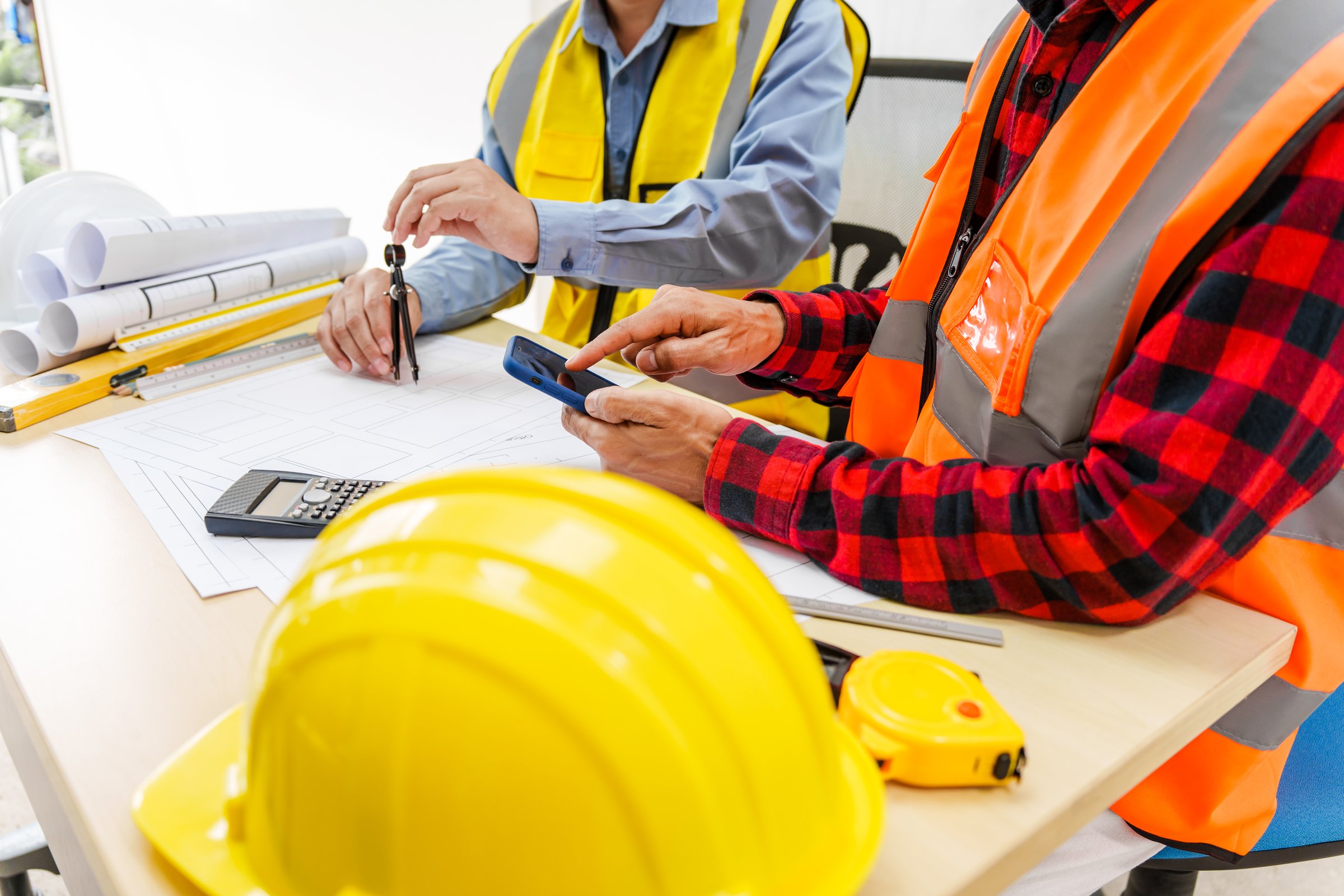 Two men, including a male Asian construction contractor engineer, discuss structural plans and engineering concrete columns and beams of a new house at a desk with a building tower model.