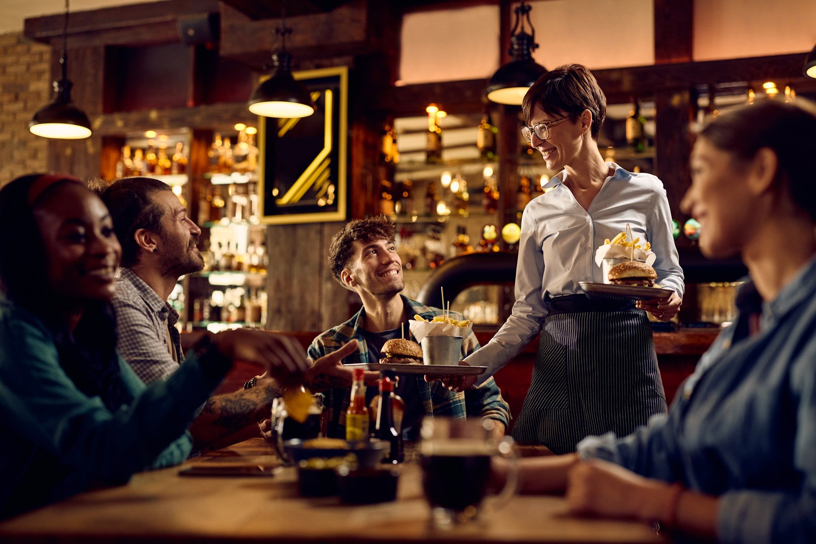 Happy waitress serving hamburger with French fries to group of guests in a bar.