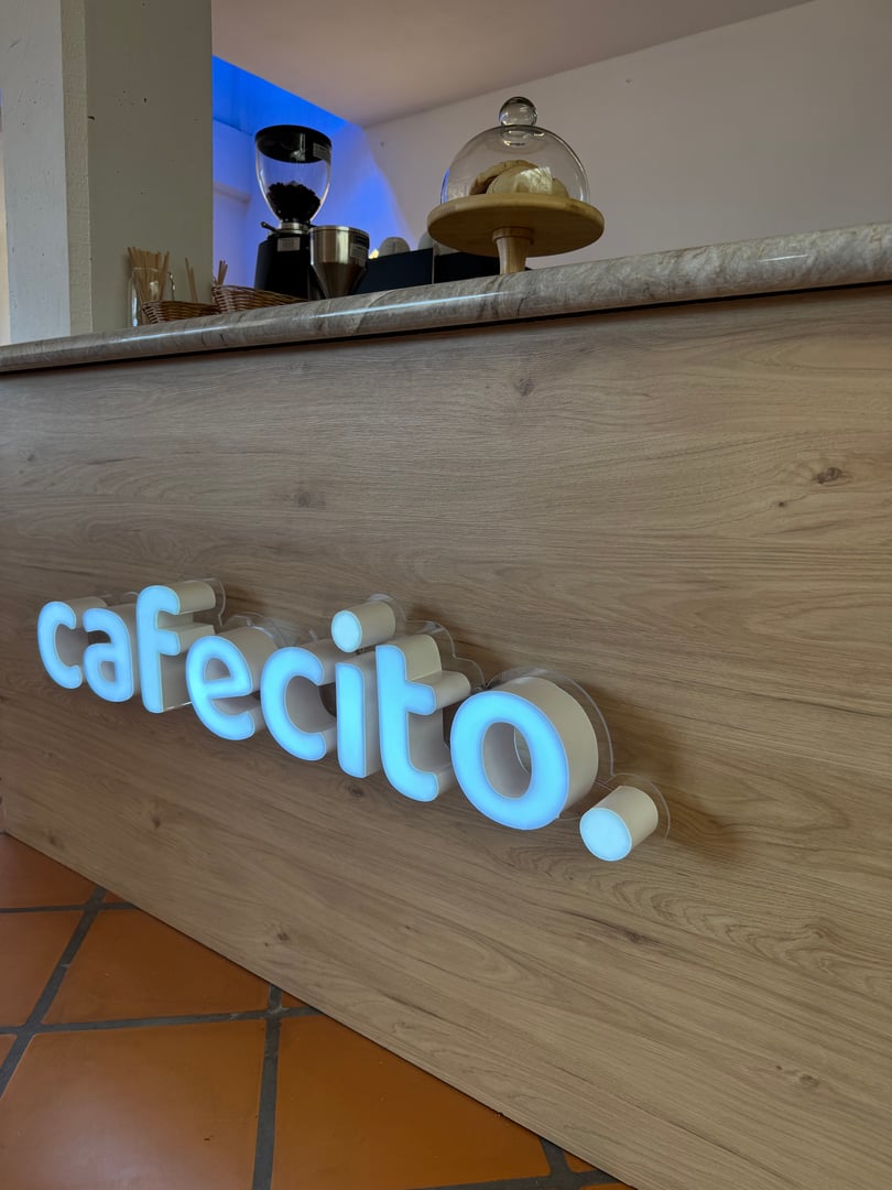 Wooden cafe counter with glowing "cafecito" sign, showcasing a cake stand and coffee grinder in the background.