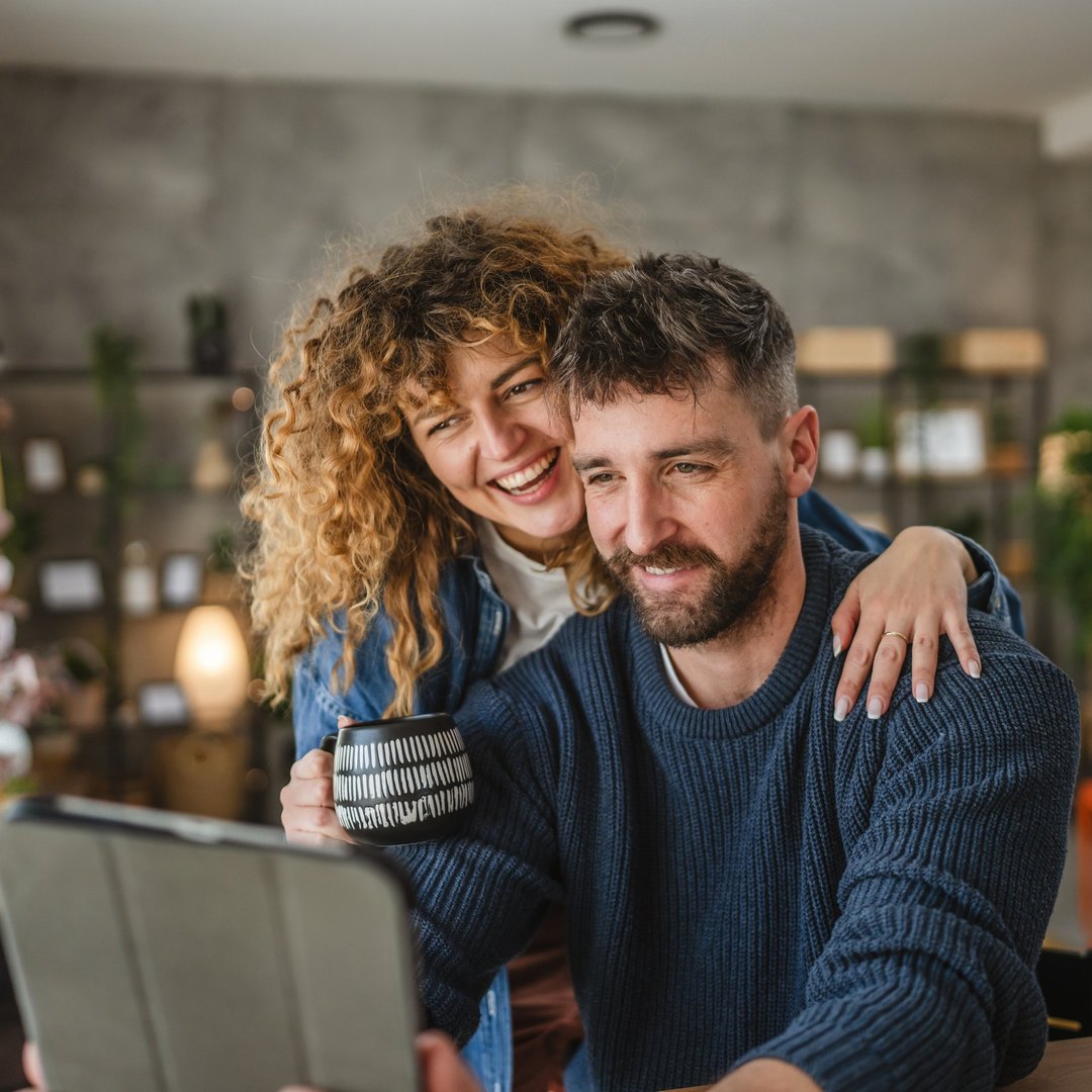 A happy adult couple sit in a cozy home setting, joyfully look at a digital tablet, highlighting themes of togetherness, modern technology, and leisure time in a comfortable environment