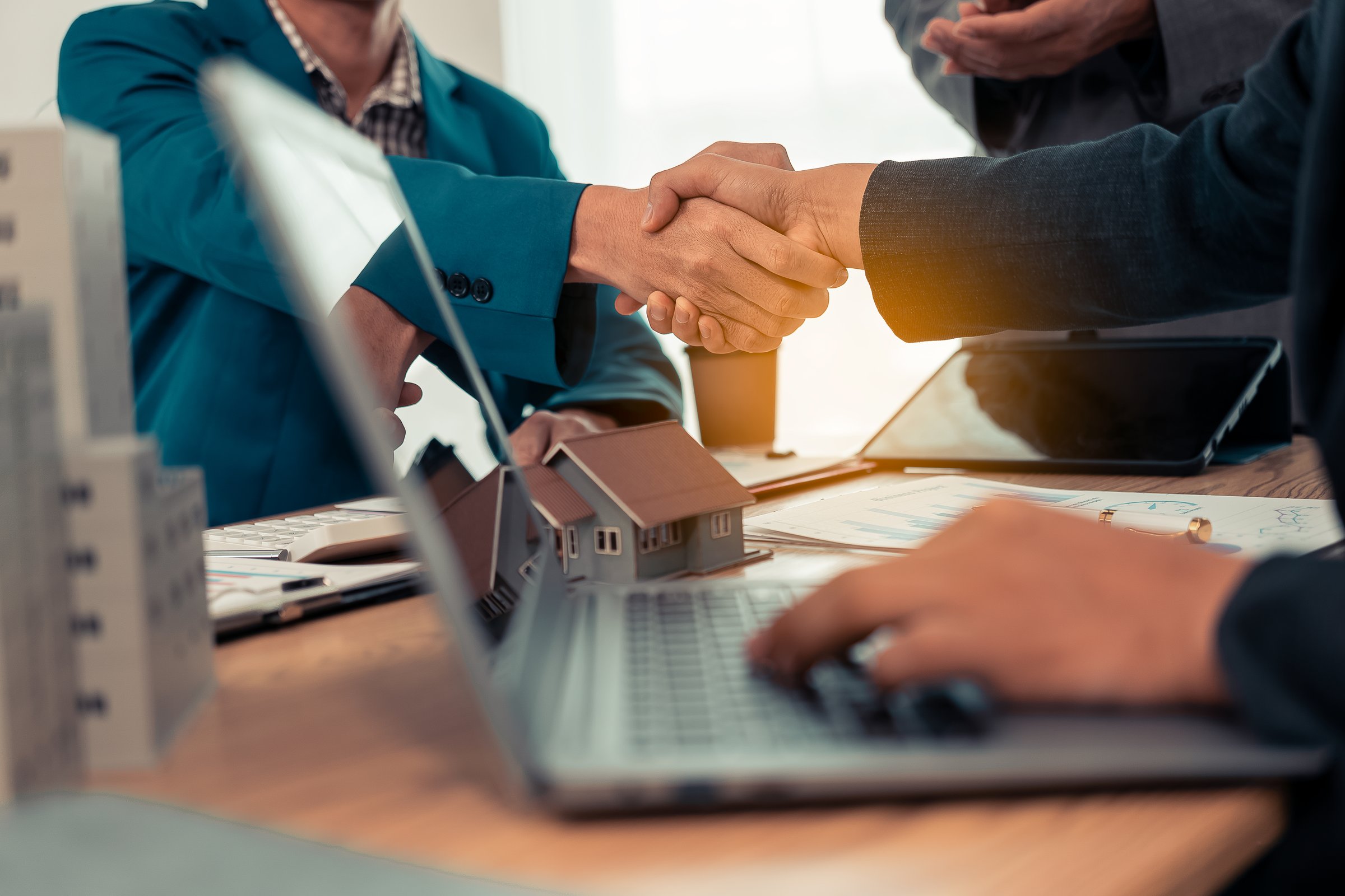 Close-up of a handshake between a real estate agent and a businessman with a small house plan in the background. Real estate, housing, transactions, contract concepts.