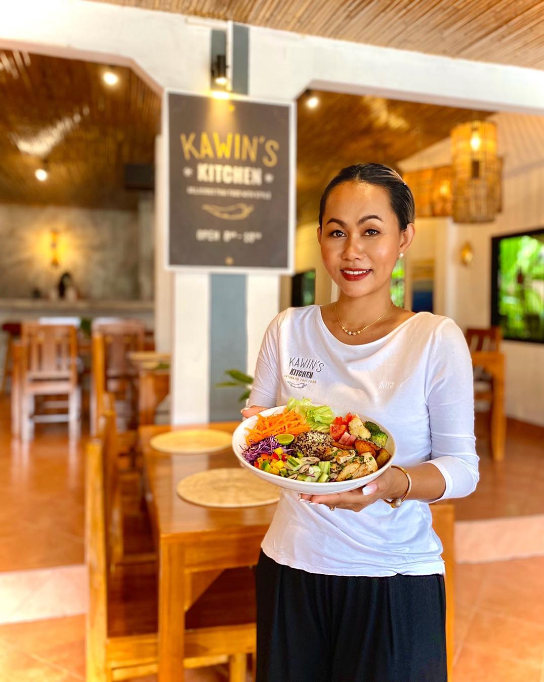 Woman holding a colorful salad bowl in a restaurant with a sign that reads Kawins Kitchen in the background.