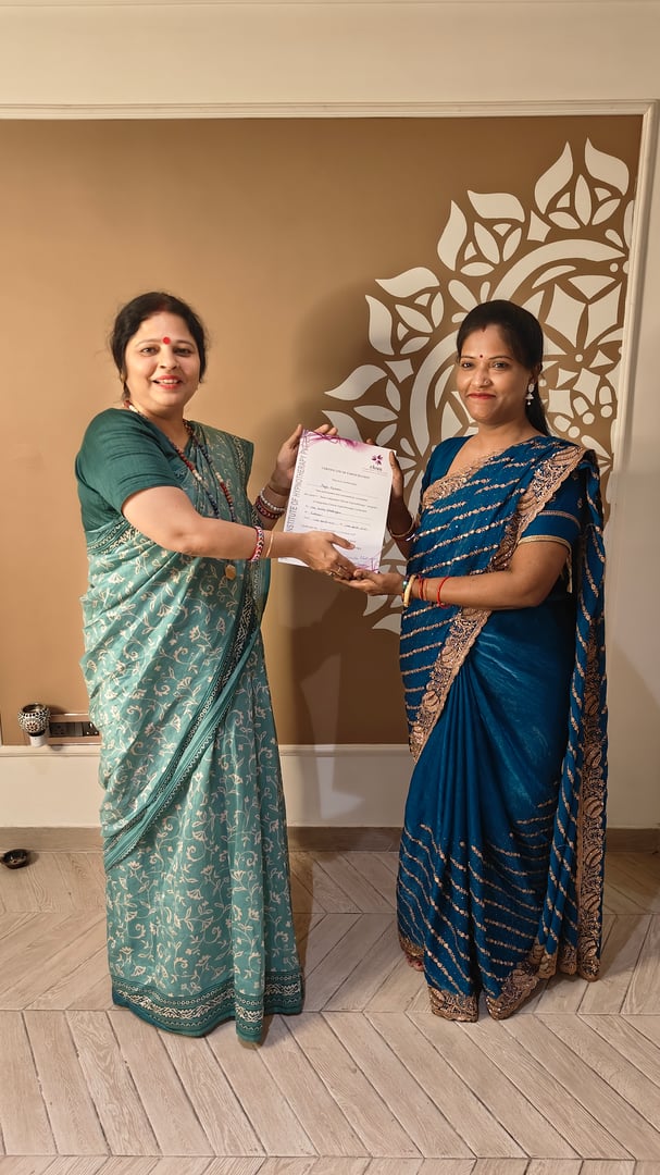 Two women in traditional attire holding a certificate, standing in front of a decorated wall.