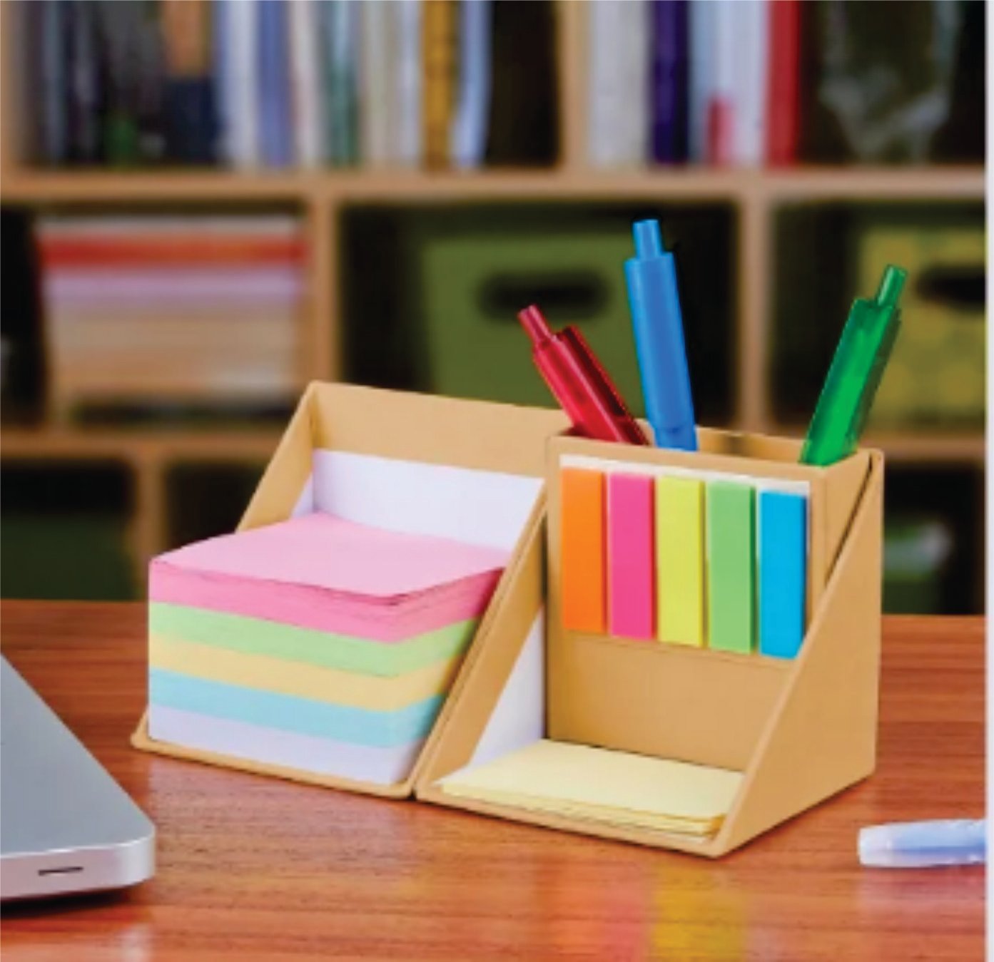Desktop organizer with sticky notes, colored pens, and note pads on a wooden desk with a blurred bookshelf background.