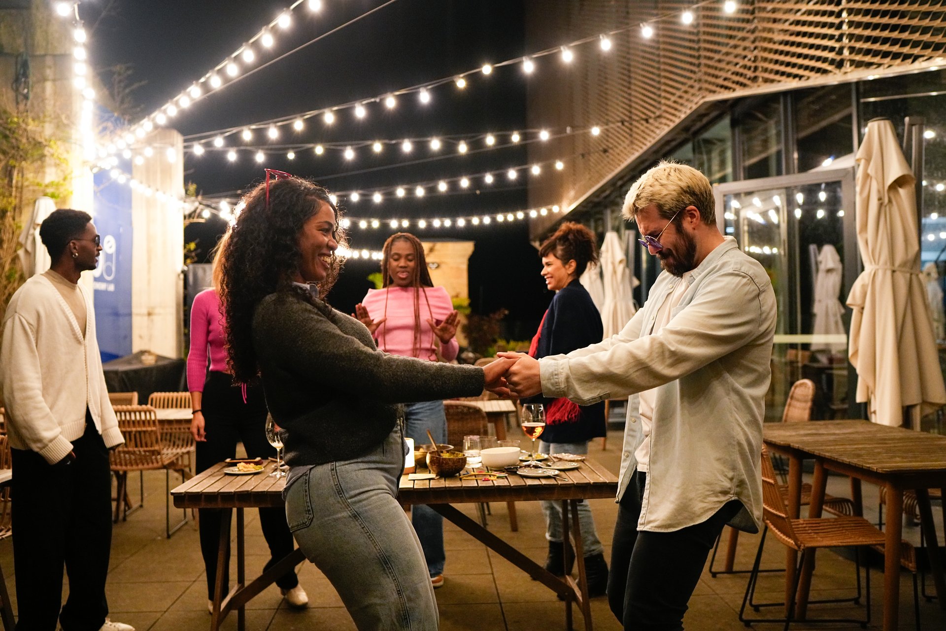 Cheerful multiethnic friends dancing and enjoying a party on a rooftop terrace with string lights at night