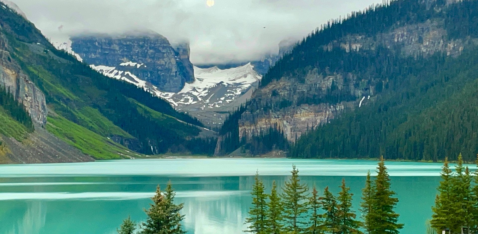 Scenic view of turquoise lake surrounded by pine trees and snow-capped mountains under a cloudy sky.