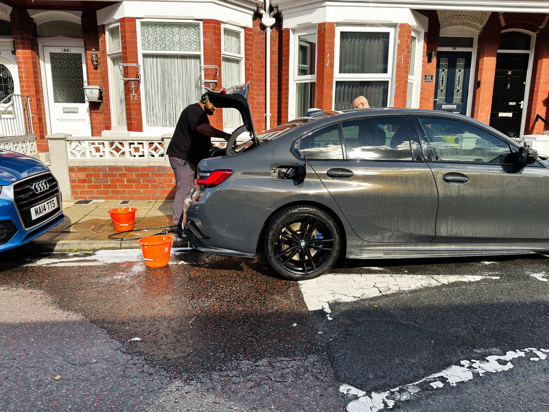 Manchester, UK - September 23, 2025: A man washes a gray BMW sedan on a Manchester street, with orange buckets nearby and a blue Audi parked.