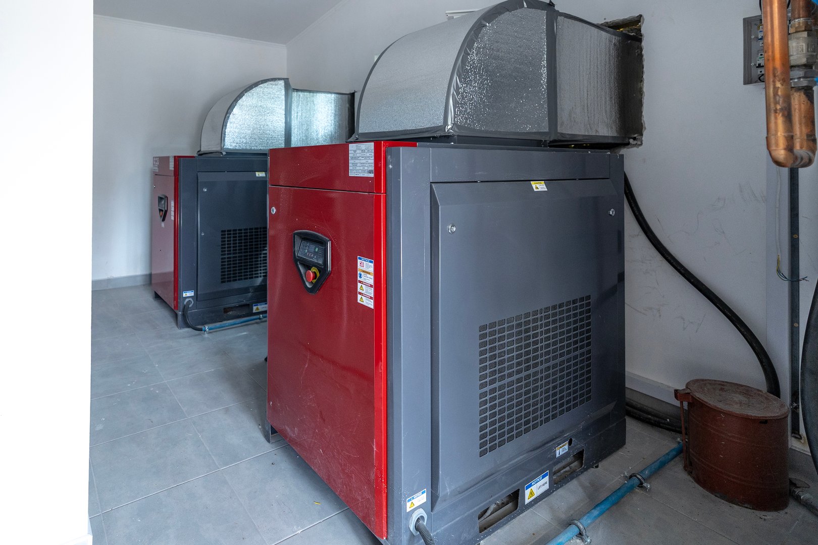 Two large industrial screw compressors, capable of 3220 liters per minute, stand in a utility room with insulated ductwork overhead, showcasing industrial machinery.