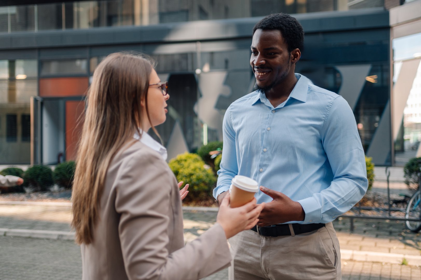 Two young professionals are standing outside their office building, engaging in a friendly conversation while holding coffee cups