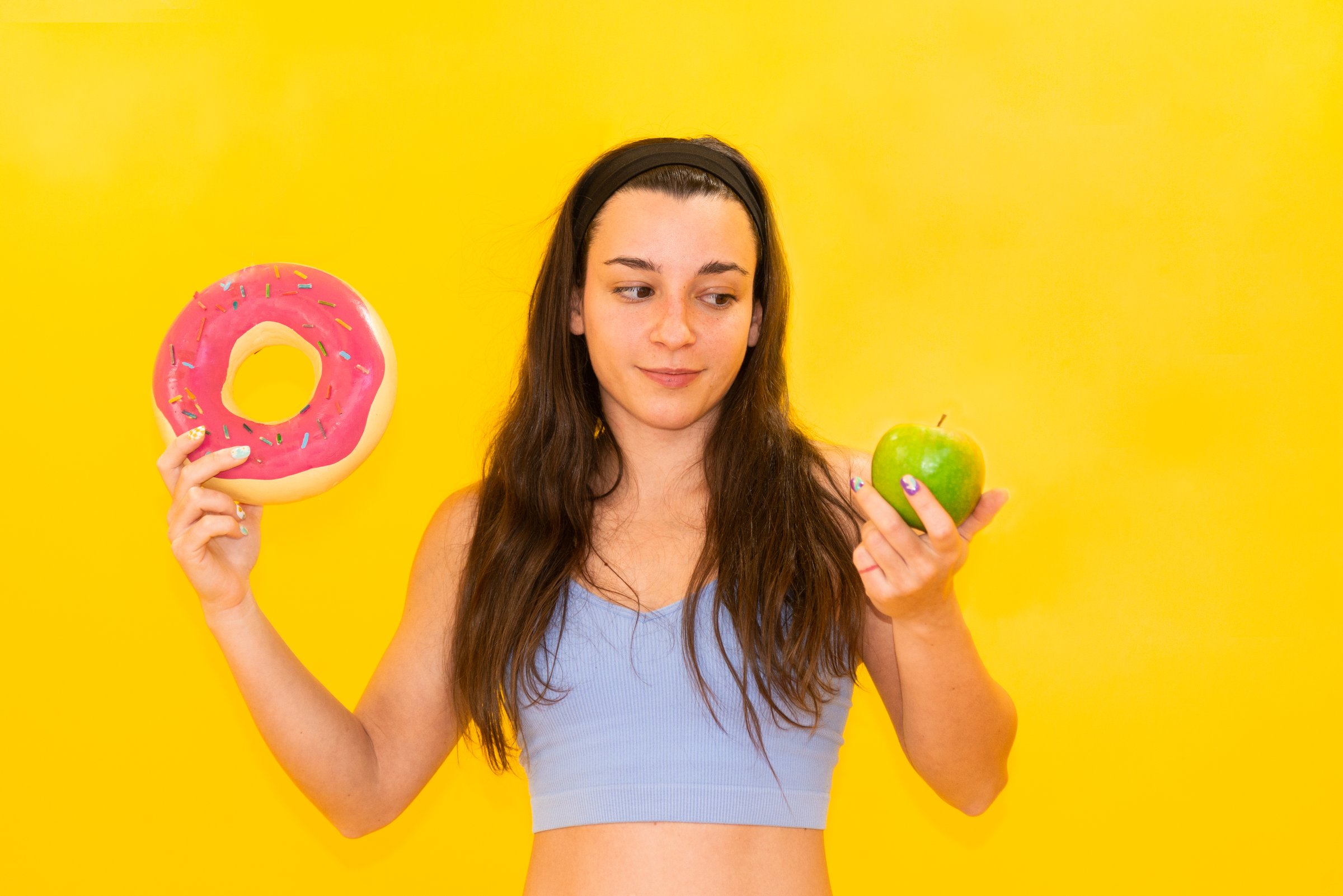 A woman holds a pink glazed doughnut in one hand and a green apple in the other, making a choice against a vibrant yellow background.