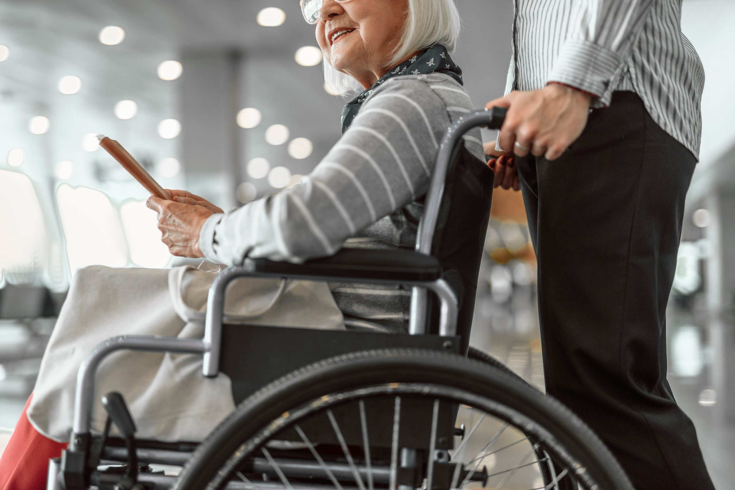 Airport female officer helping for old female on disabled carriage while waiting in lobby. Mature lady in glasses holding ticket and smiling