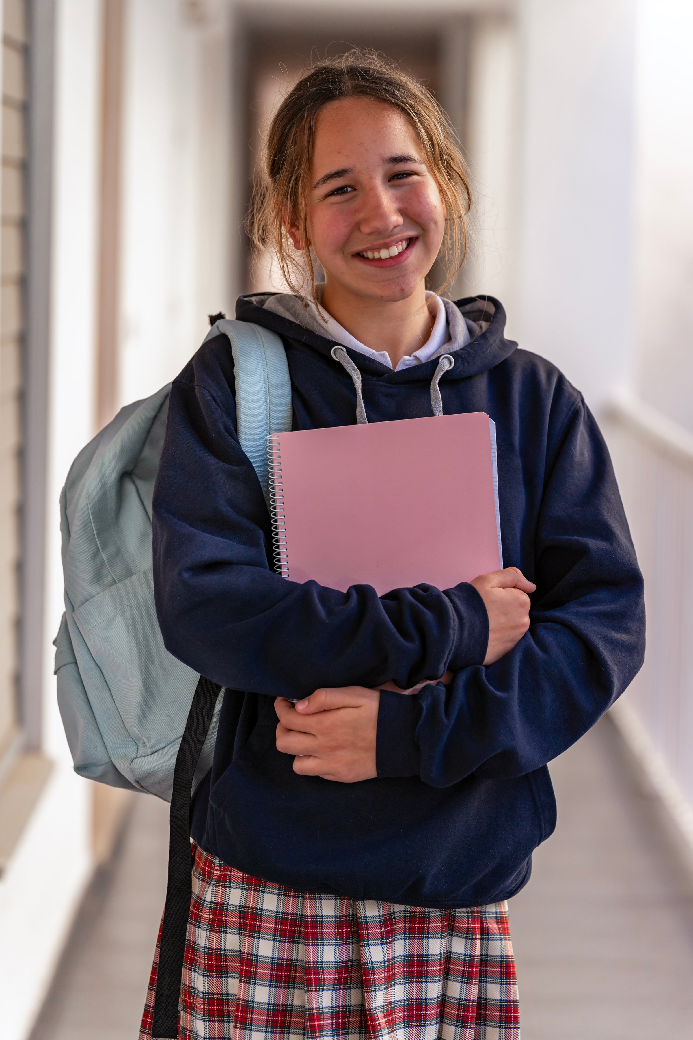 Portrait of a smiling student girl with backpack wearing uniform and holding books in a high school hallway.
