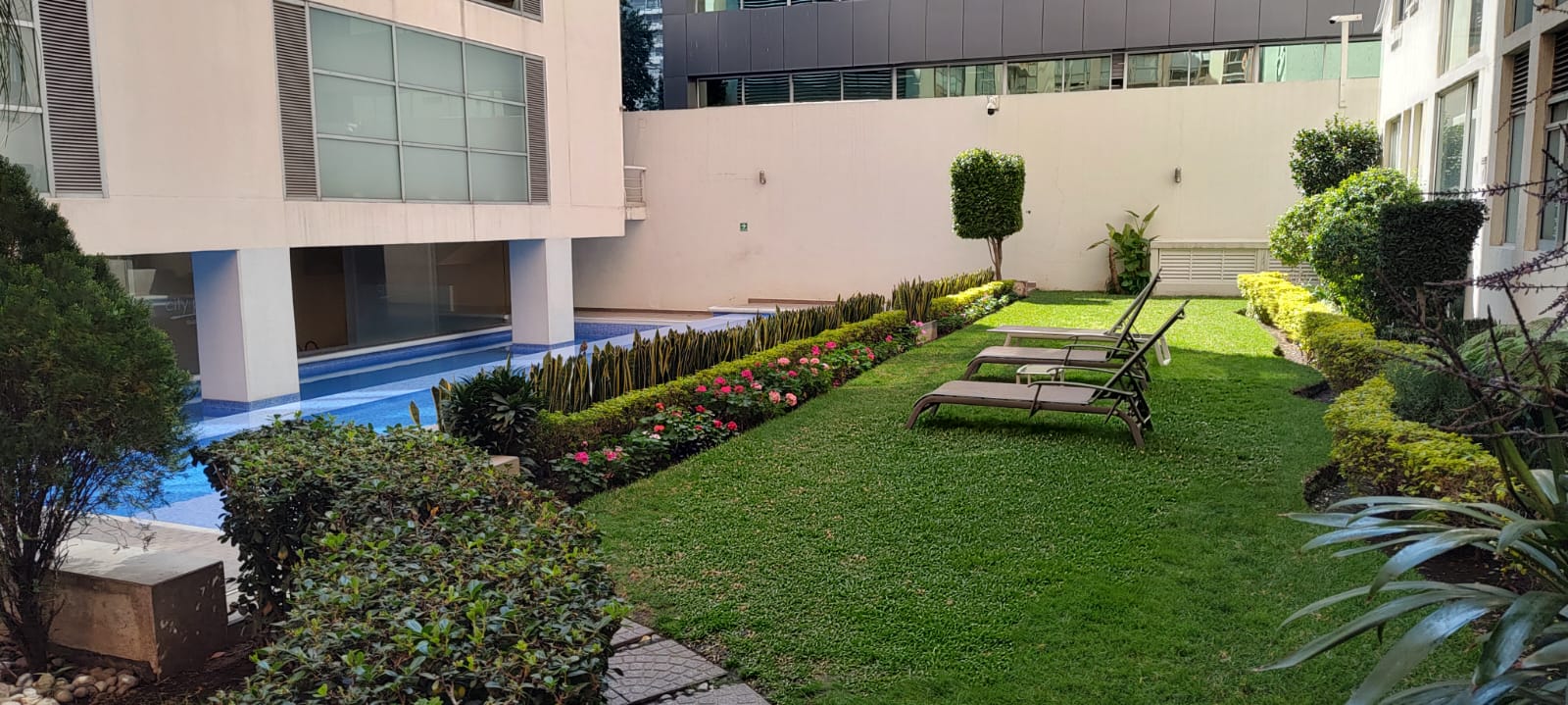 Apartment complex courtyard with a pool, lounge chairs, trimmed bushes, and colorful flowers.