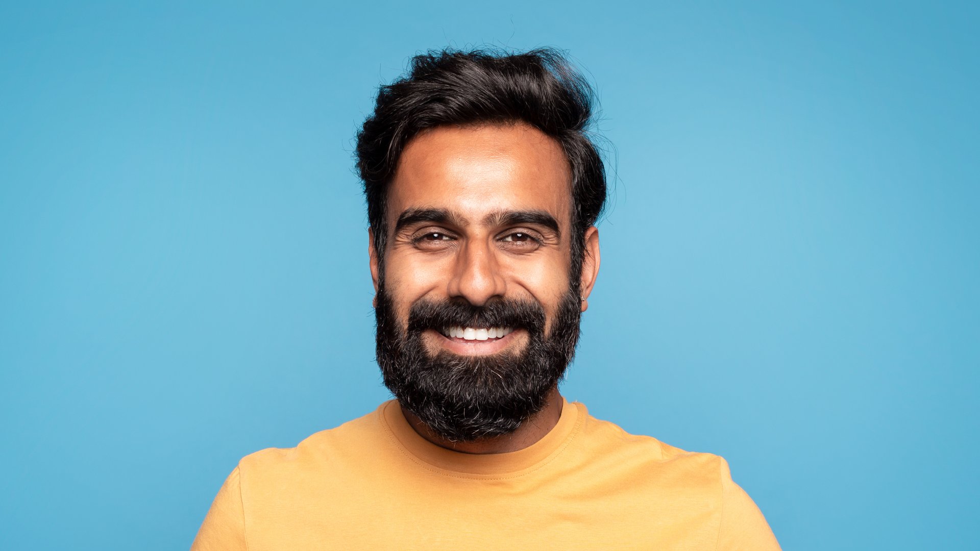Closeup portrait of happy middle aged indian man in casual cheerfully smiling at camera posing over blue studio background, showing white teeth, panorama. Male portrait concept