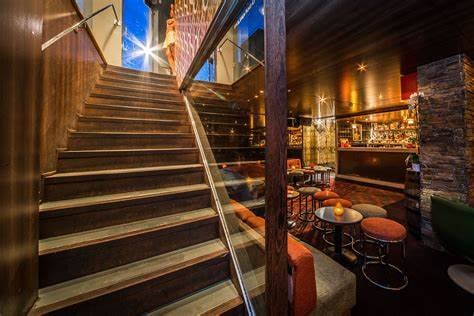 Stylish bar interior with wooden stairs, cushioned seating, ambient lighting, and a stocked bar in a cozy atmosphere.