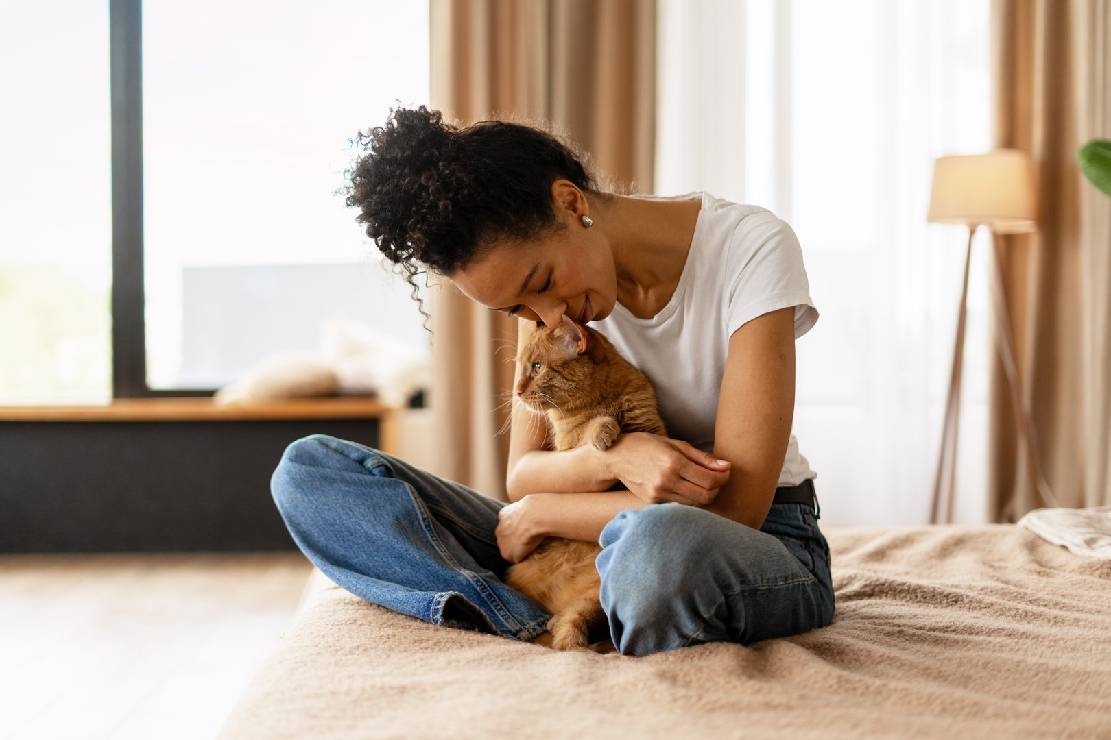 Young woman sitting on bed, embracing and showing affection to her ginger cat in a cozy home environment
