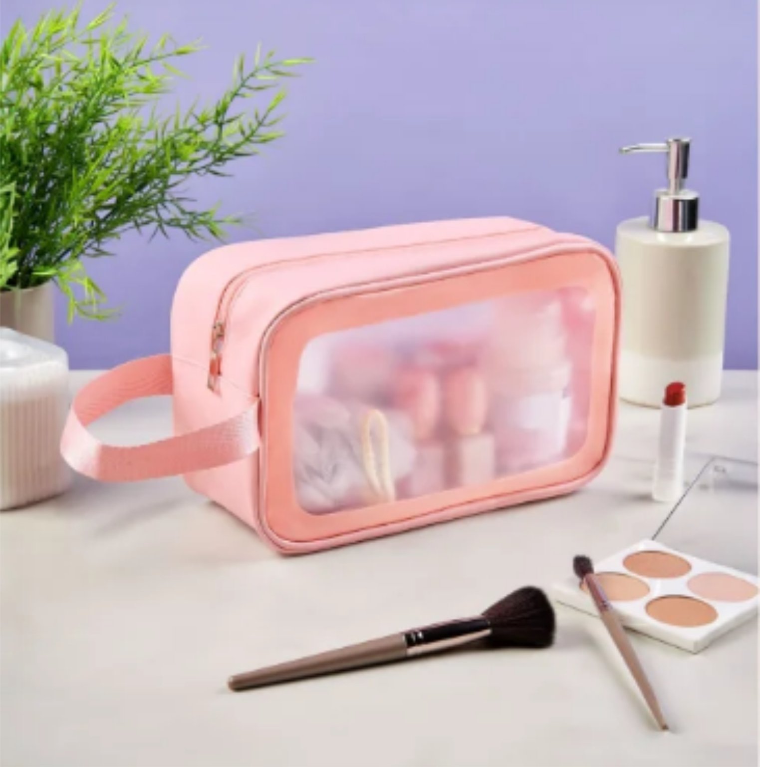 Pink cosmetic bag with transparent front on a vanity surrounded by makeup items, a plant, and a soap dispenser.